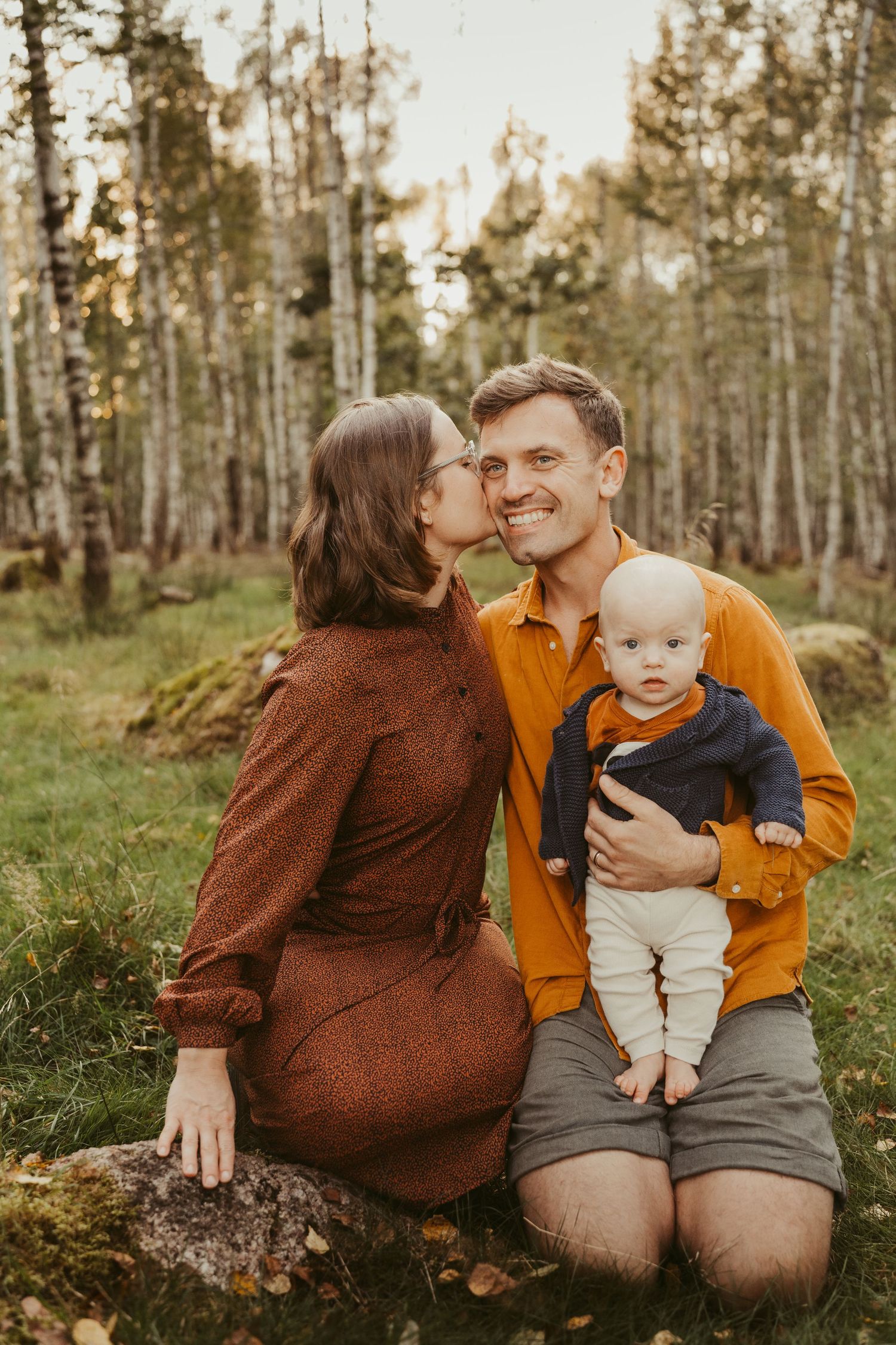 A family poses together in a forest setting wearing brown and orange autumn clothing.