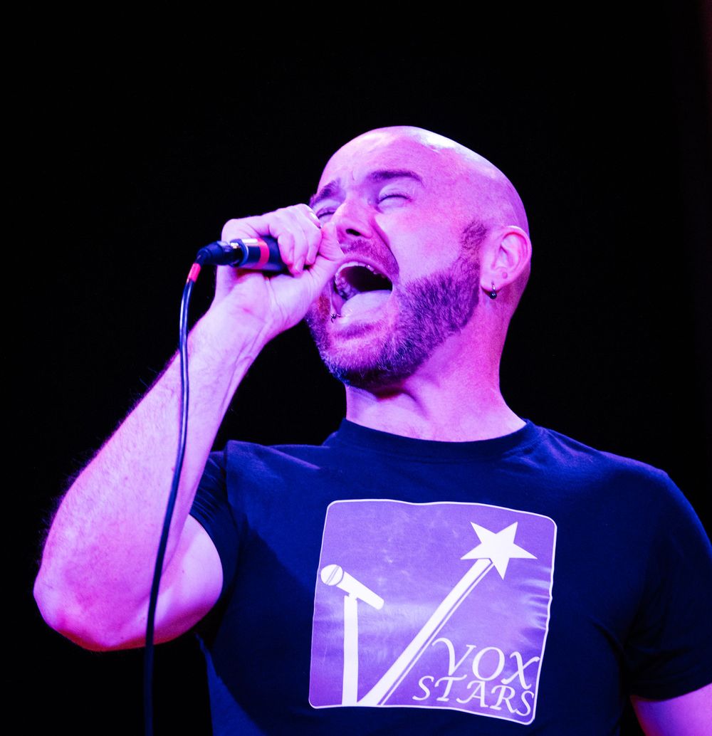 A performer sings passionately into a microphone under purple stage lighting.