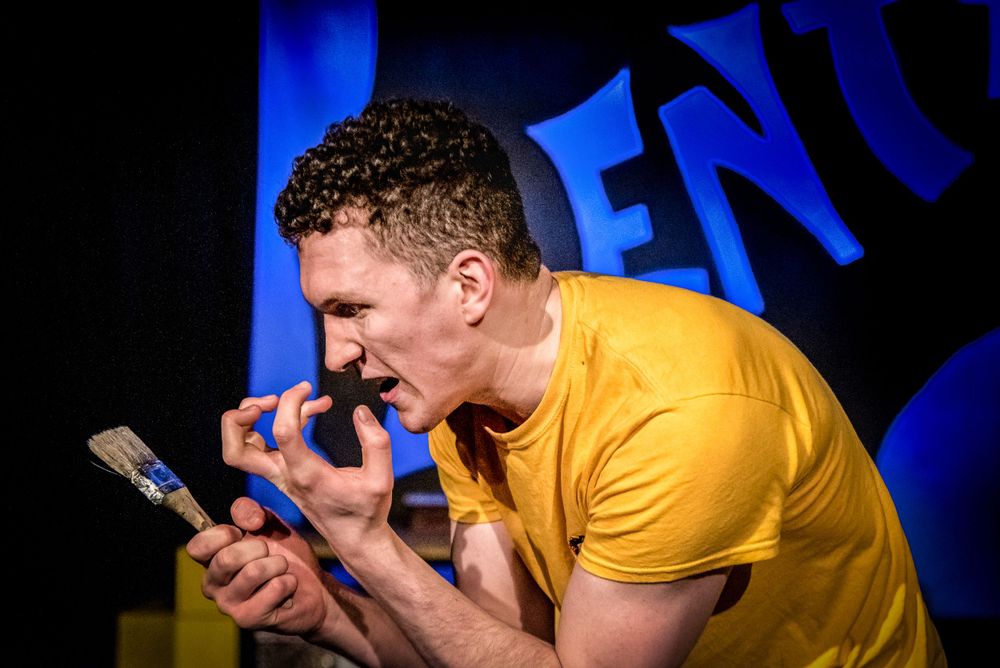 A performer in a yellow t-shirt reacts expressively while looking at a mobile phone on stage.