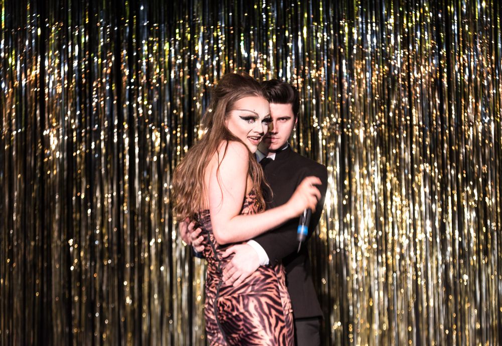 A couple embracing in formal attire against a gold sparkly backdrop.