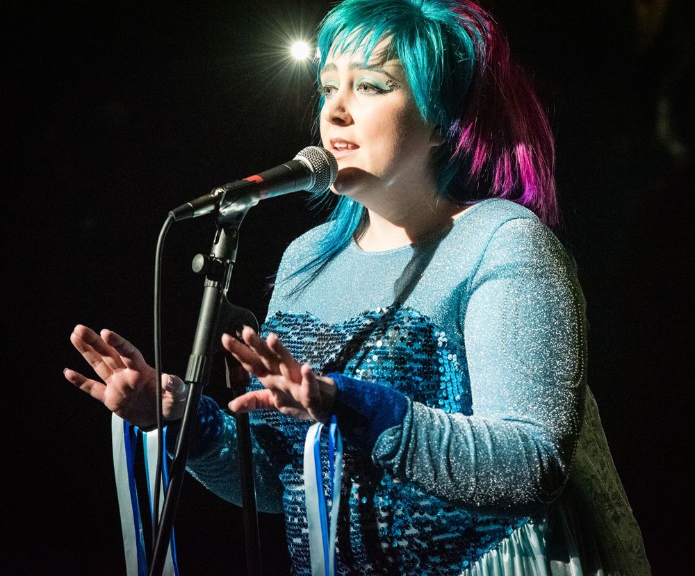 Singer with teal hair performing on stage with microphone in sparkly outfit under dramatic lighting.