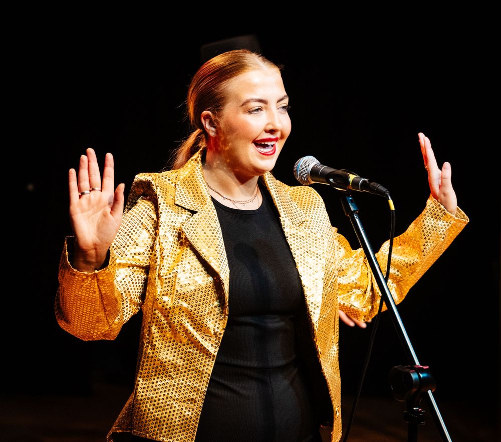 A performer in a gold sequined jacket sings enthusiastically on stage.