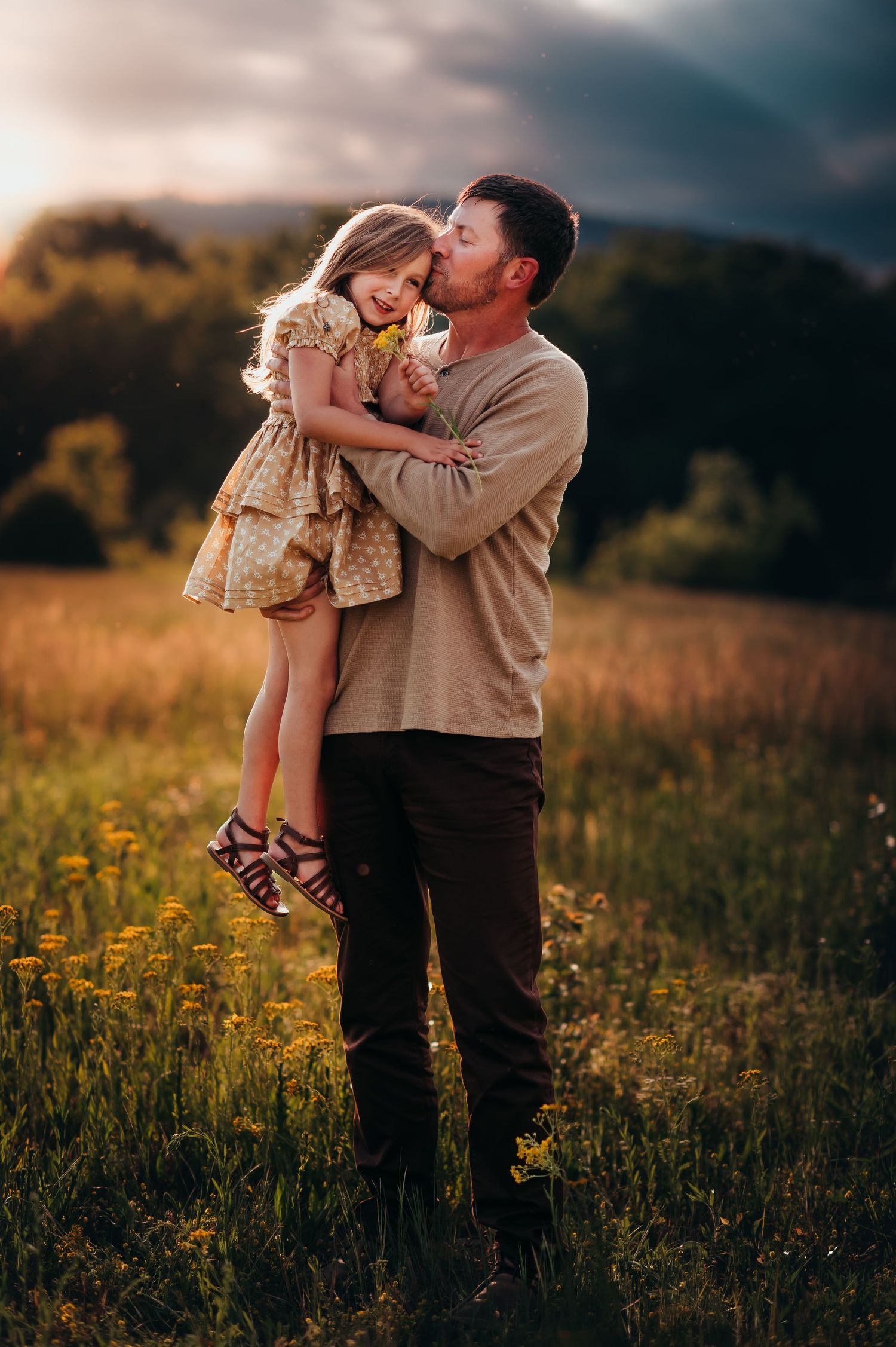 Father holds up his daughter and kisses her cheek at sunset while she holds yellow flowers