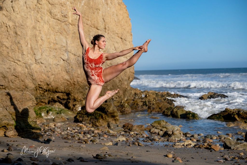 Dancer leaps gracefully on beach with cliffs and ocean waves in background.