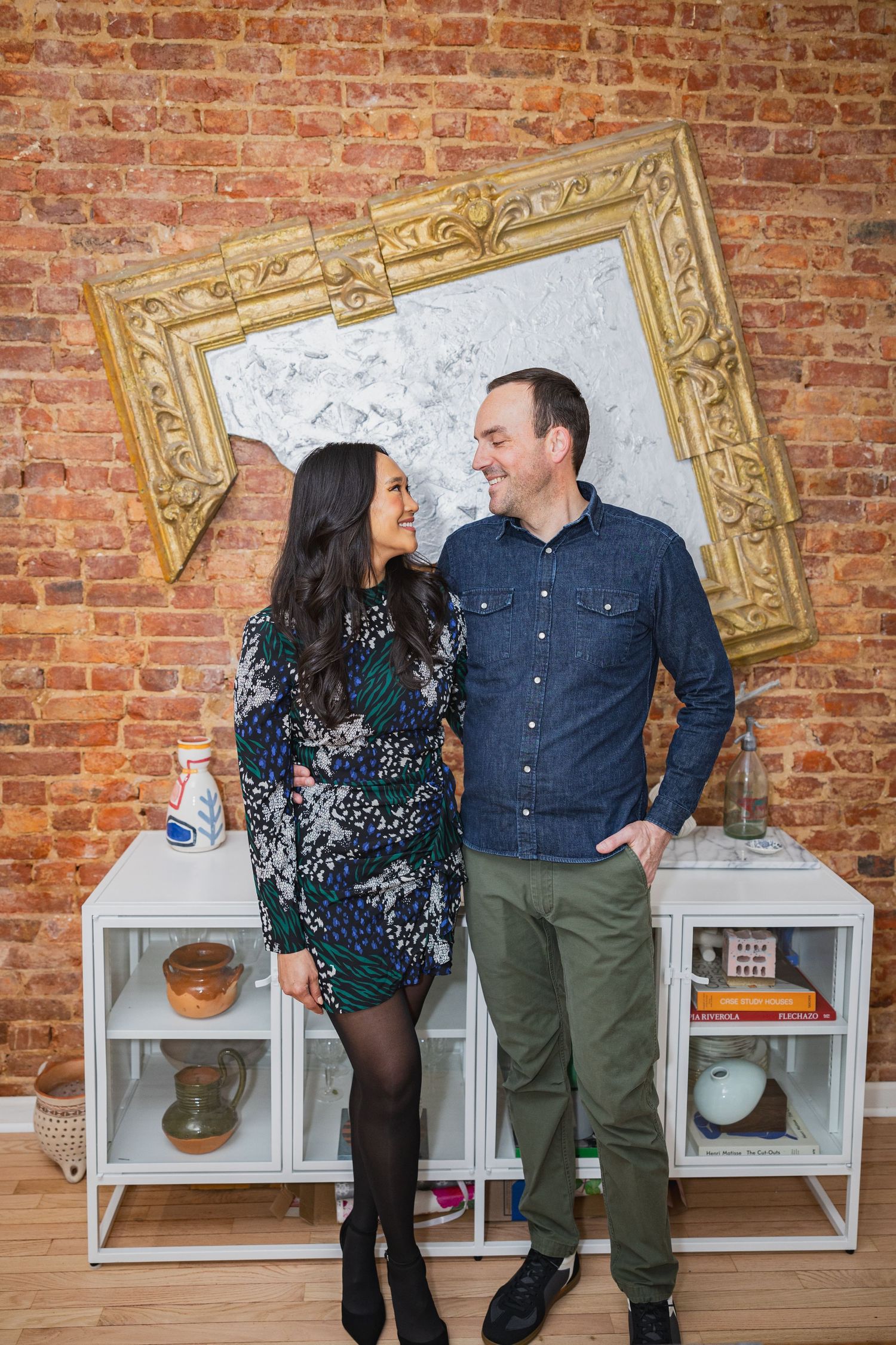 A couple smiles at each other in front of a brick wall with a decorative gold frame.