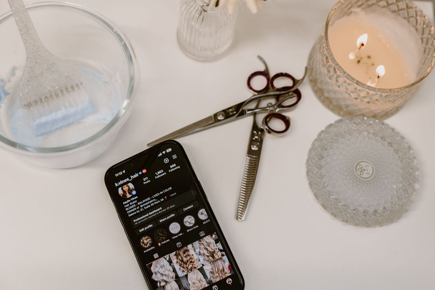 Styling tools including scissors and a phone displaying photos on a white surface with candles.