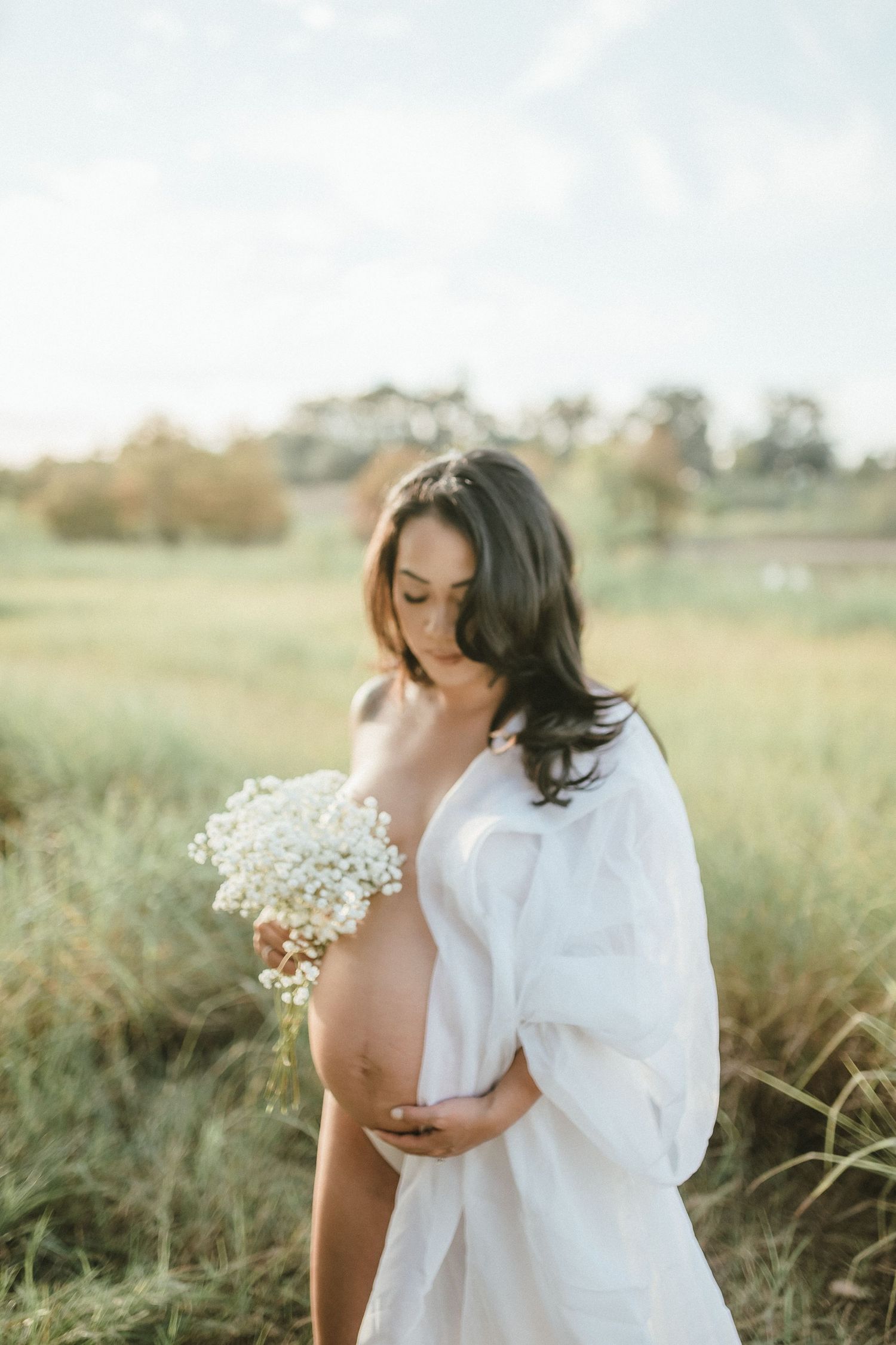 Artistic maternity photo of woman in white shirt holding baby's breath flowers in sunny field.