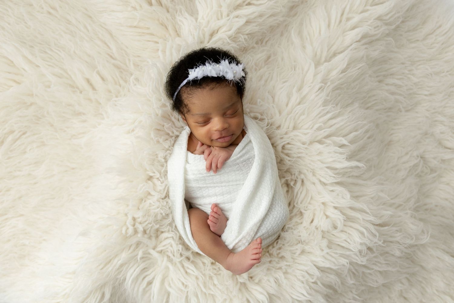 Sleeping newborn baby with headband wrapped in white against fluffy cream background.