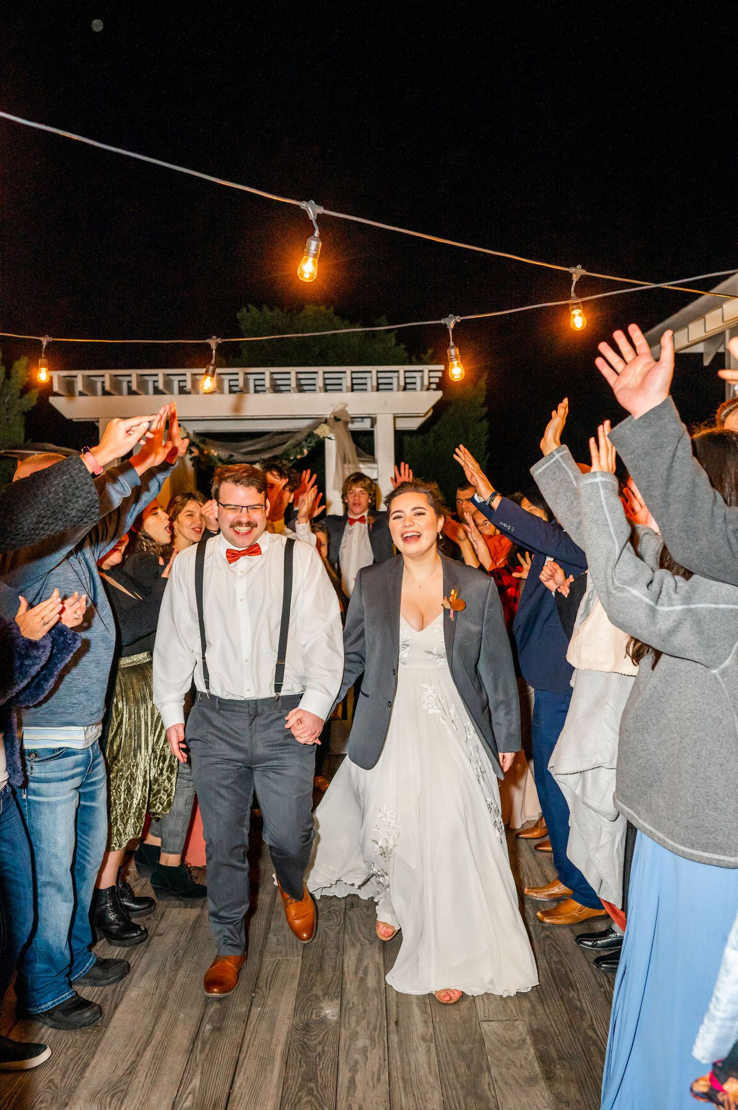 Wedding reception guests dancing and celebrating under string lights on outdoor deck at night.