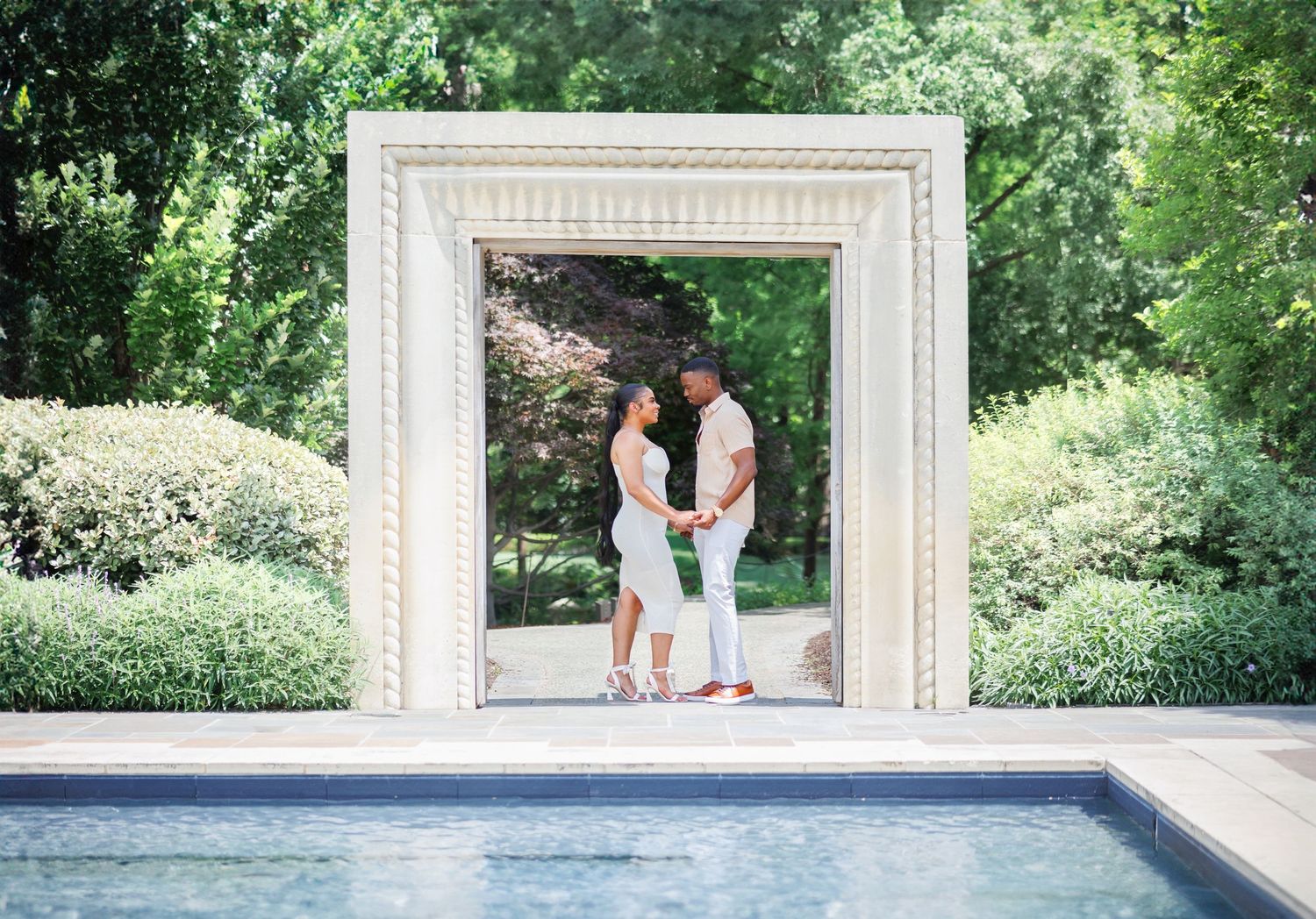 Romantic moment framed through white architectural arch near swimming pool in garden setting.