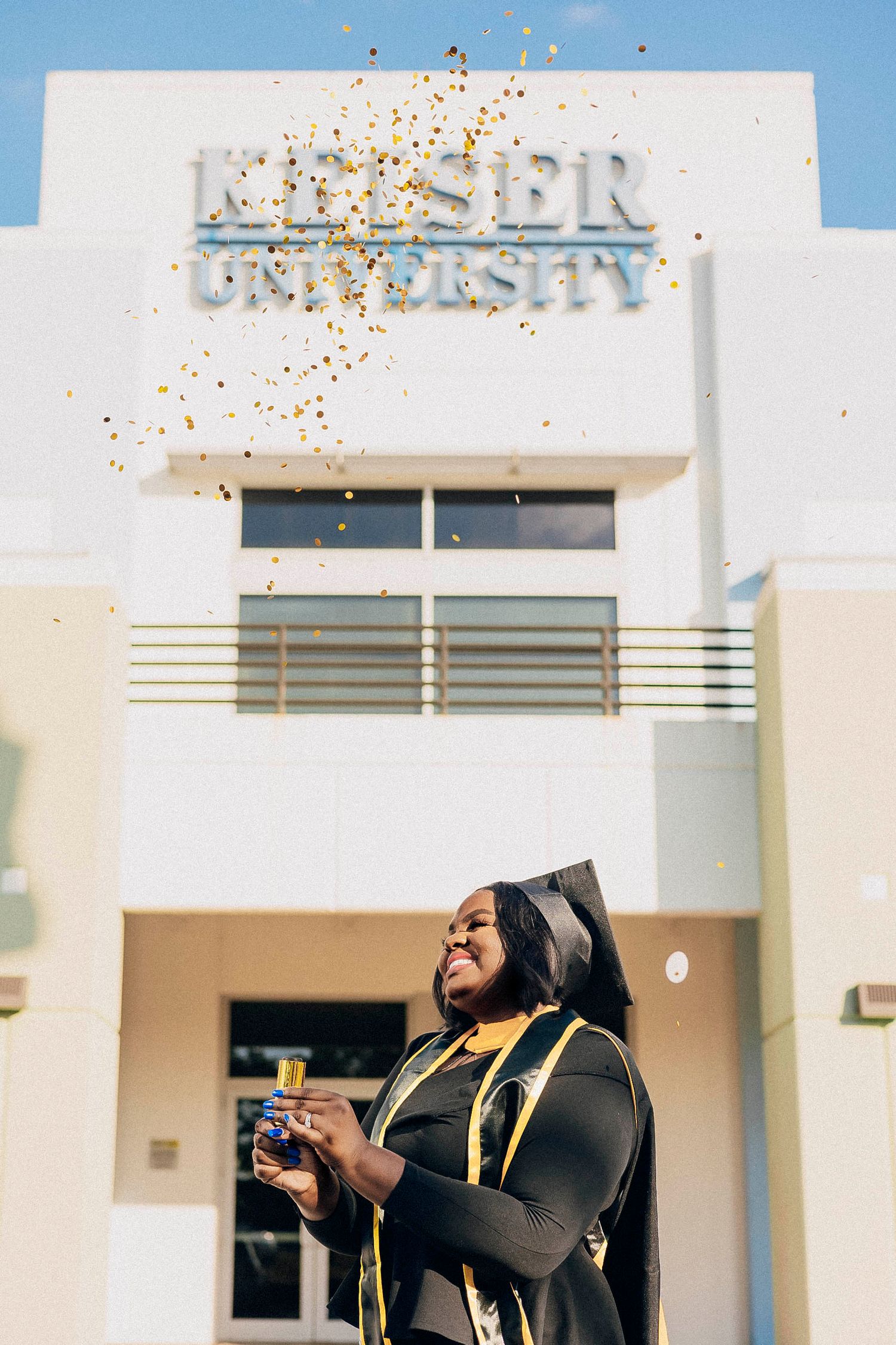 A graduate in cap and gown celebrates with gold confetti outside Keiser University building.
