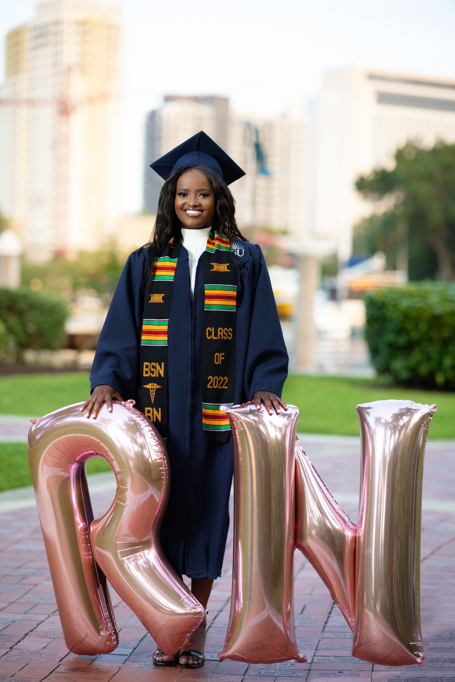 BSN RN Class of 2022 graduate poses in navy cap and gown with kente stole beside RN balloons.