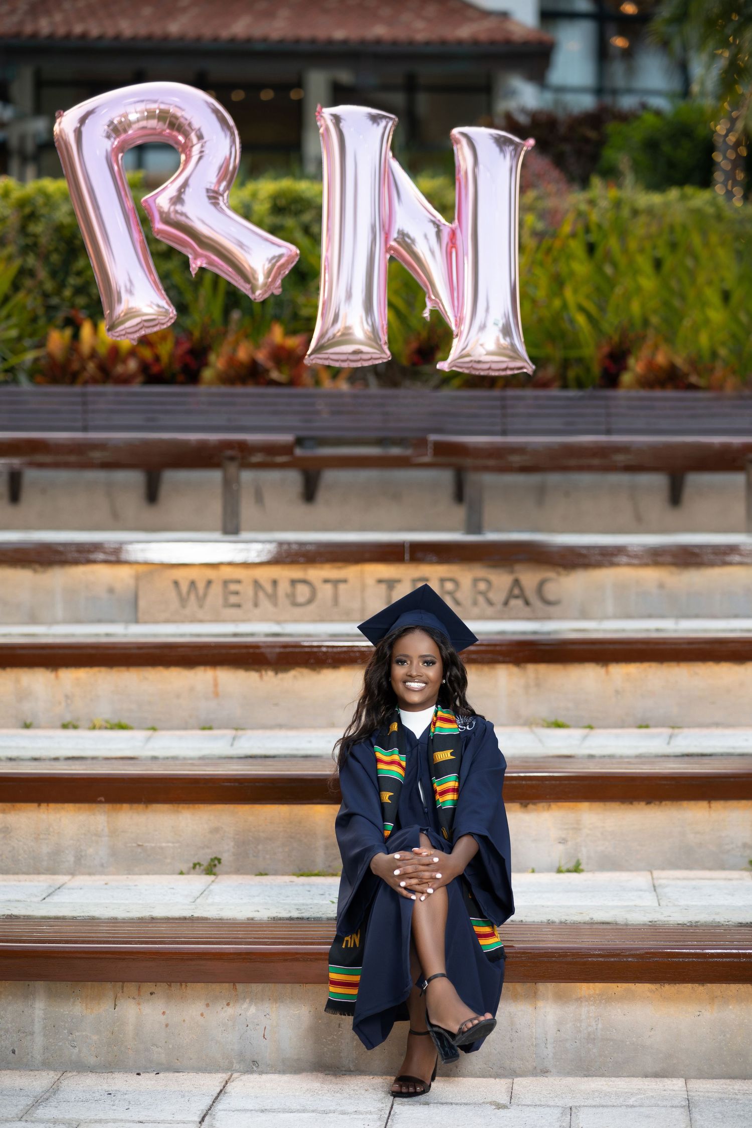 Nursing graduate in navy cap and gown sits on steps with RN balloons at Wendt Terrace.