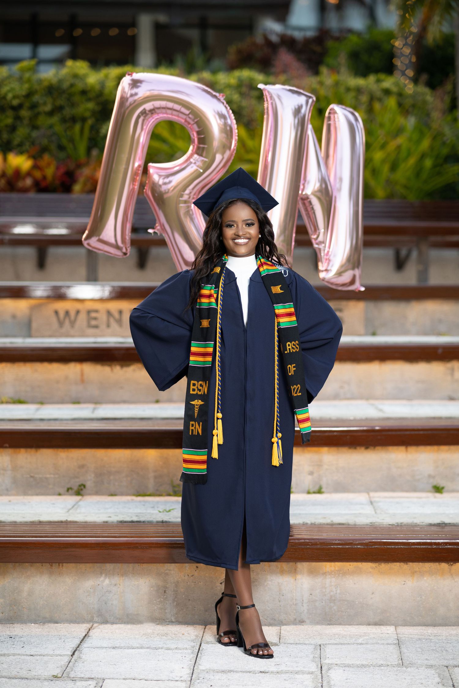 Nursing graduate in navy cap and gown with kente stole stands confidently before large RN balloons.