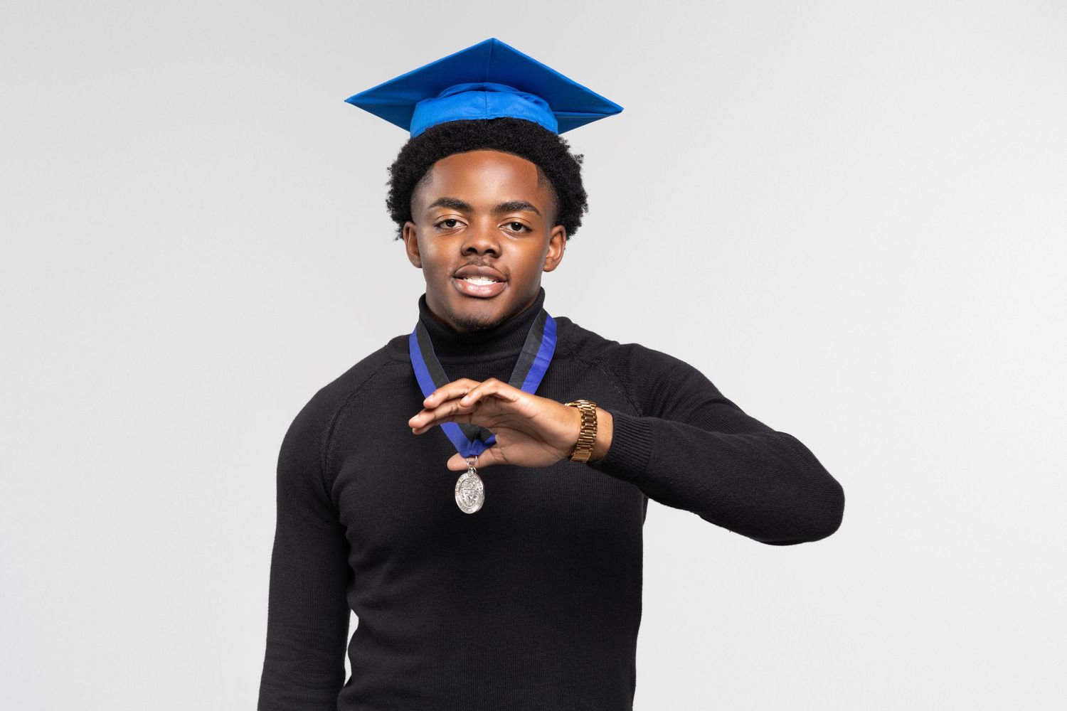 Young man in blue graduation cap and black turtleneck displays a medal while making a hand gesture.