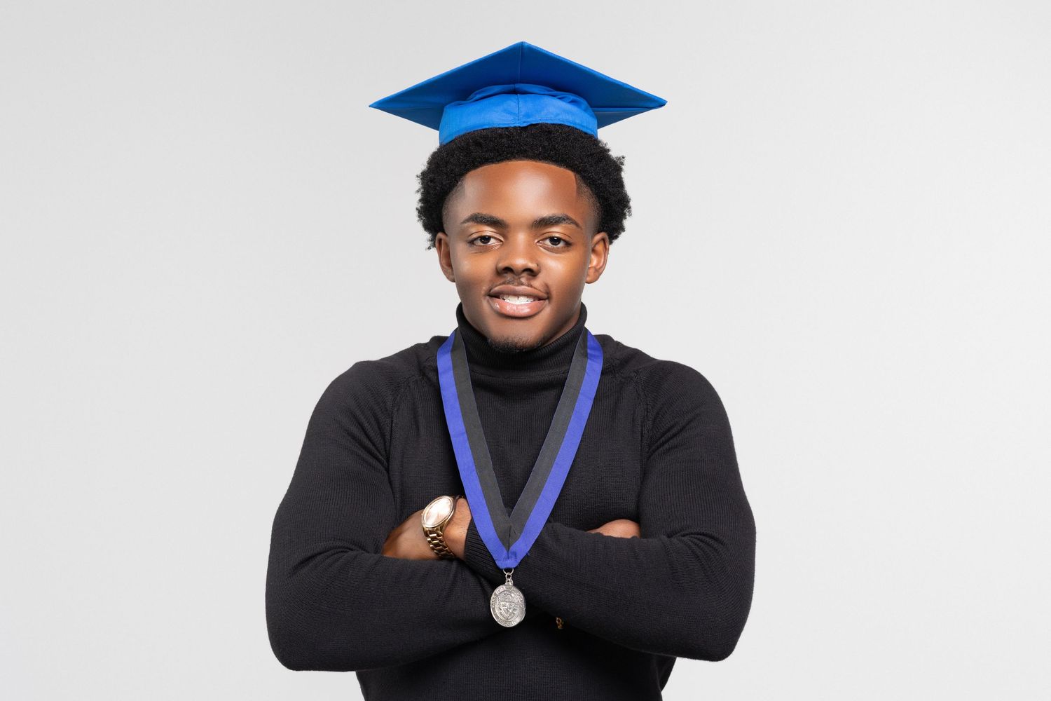Confident young male graduate in blue cap and black turtleneck stands with arms crossed wearing a medal.
