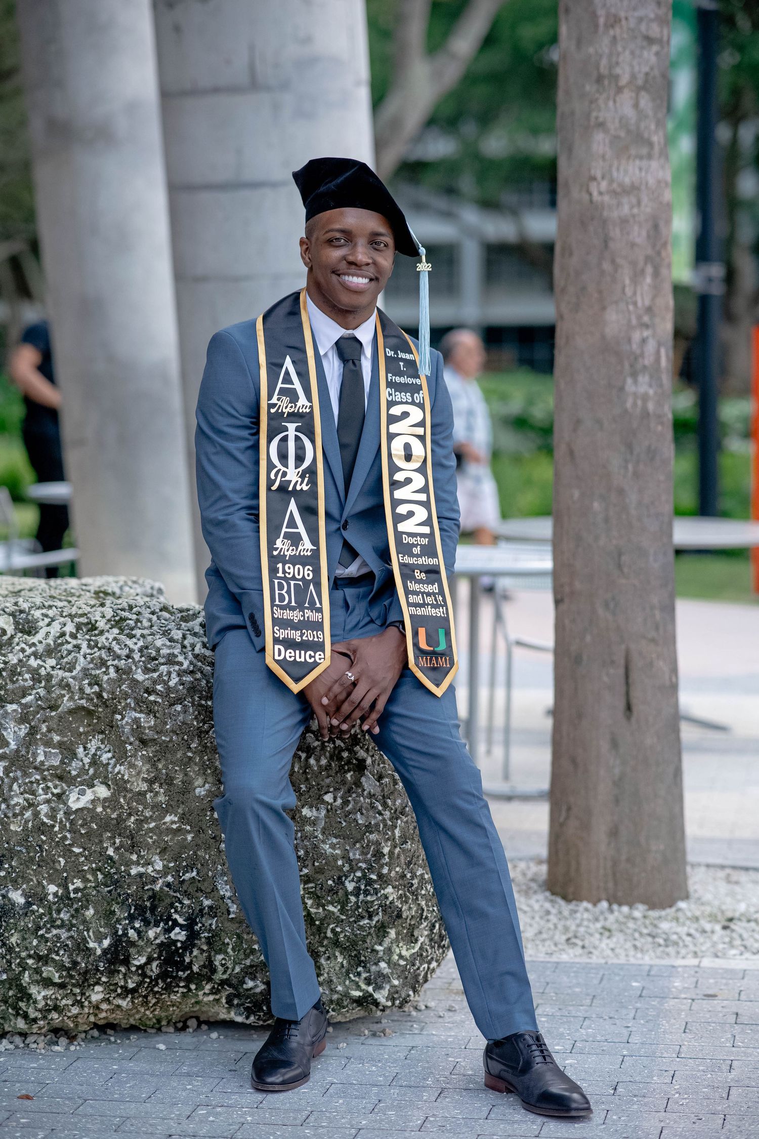 Smiling graduate in gray suit and cap leans against stone wall wearing Alpha Phi Alpha and university stoles.