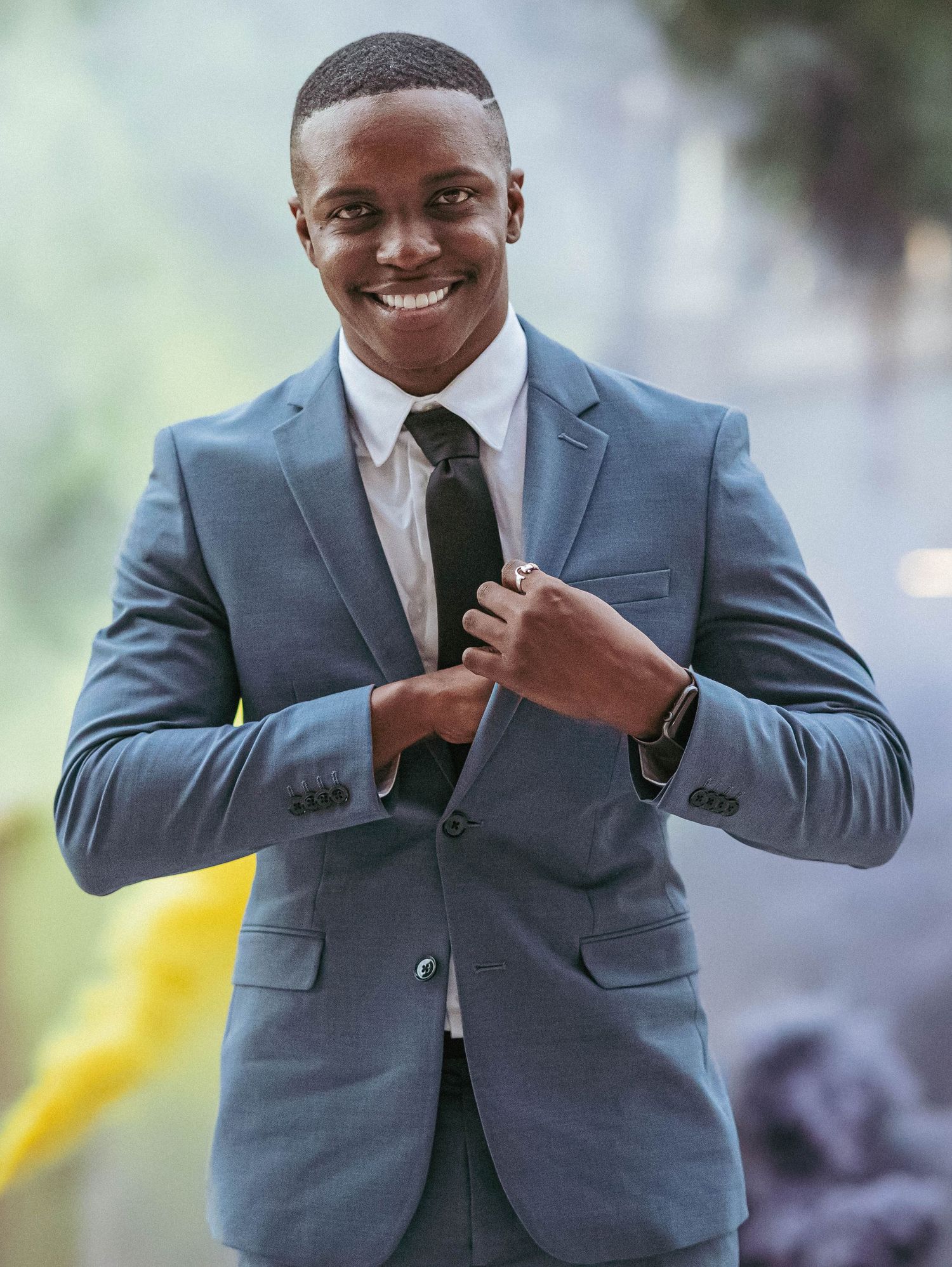 Smiling young Black man in grey suit adjusting tie for graduation celebration portrait.