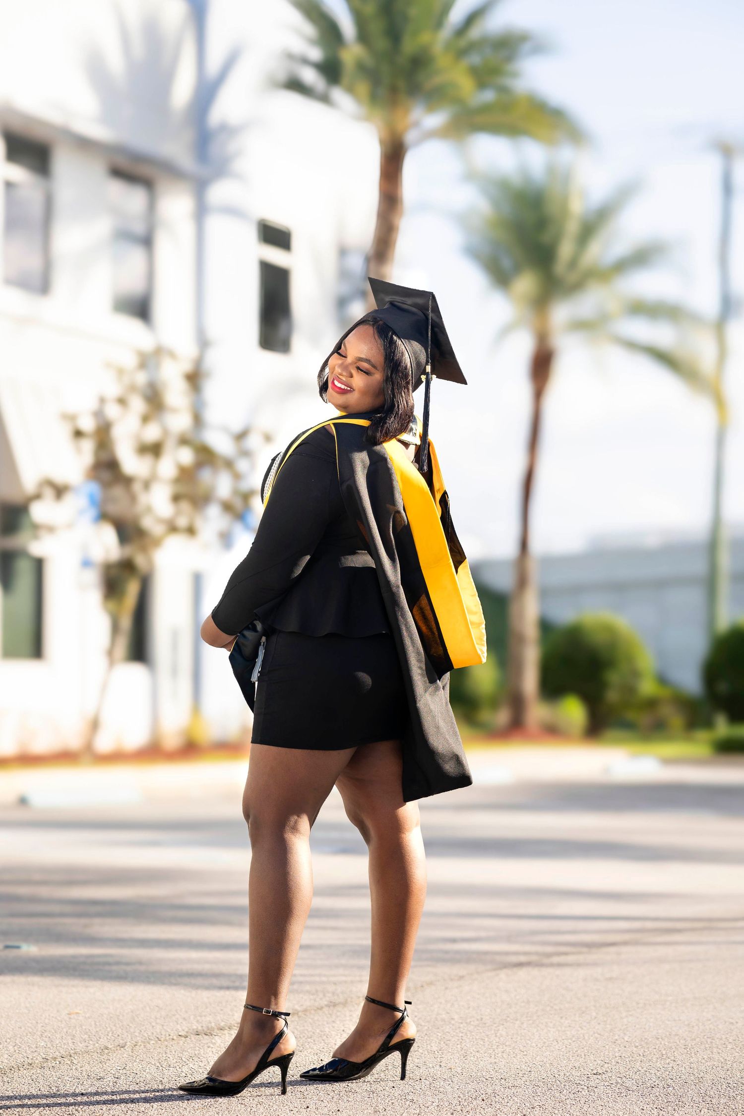 Smiling graduate in black and gold cap and gown posing on a sunny street lined with palm trees.