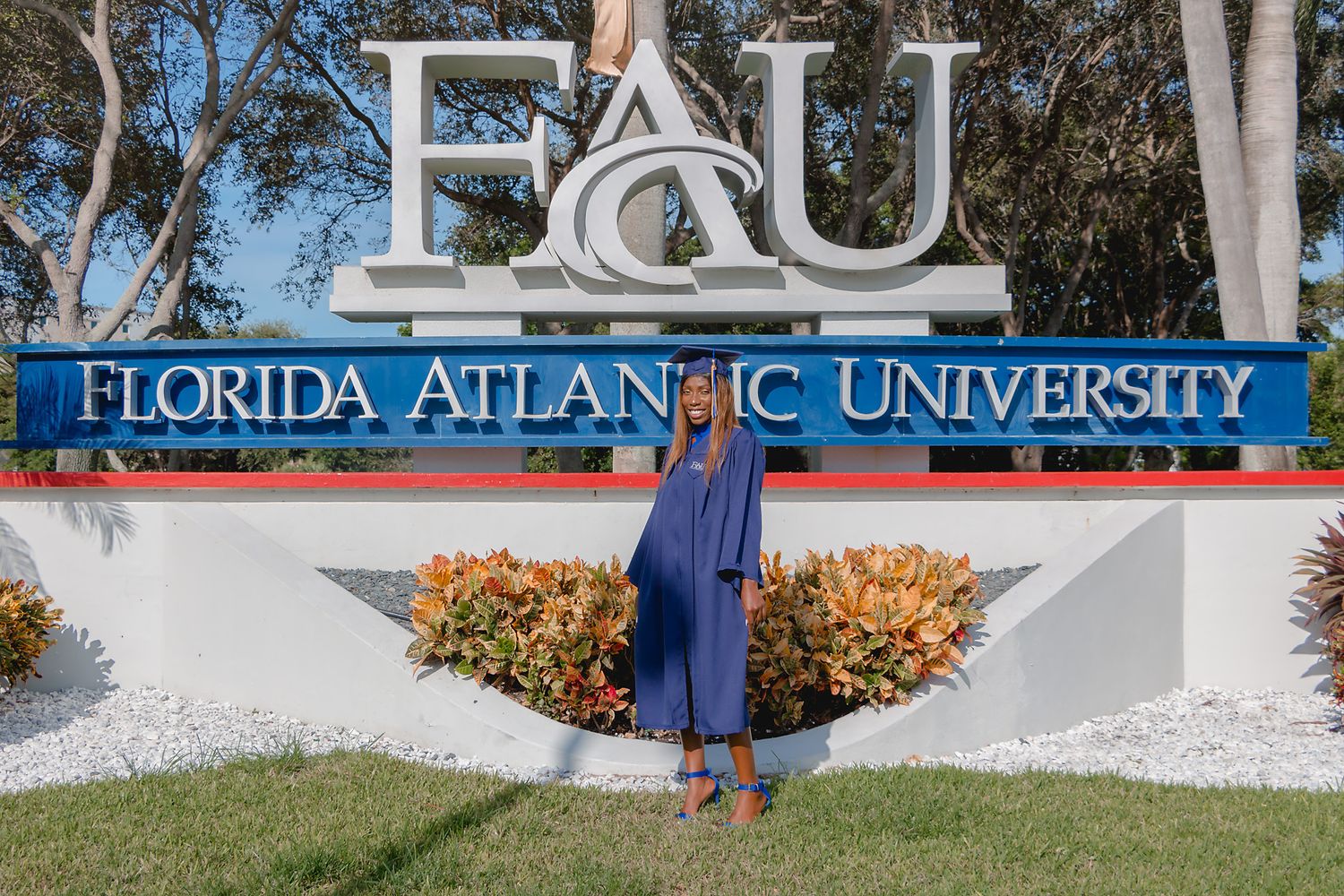 Proud graduate poses in blue cap and gown in front of Florida Atlantic University FAU sign.