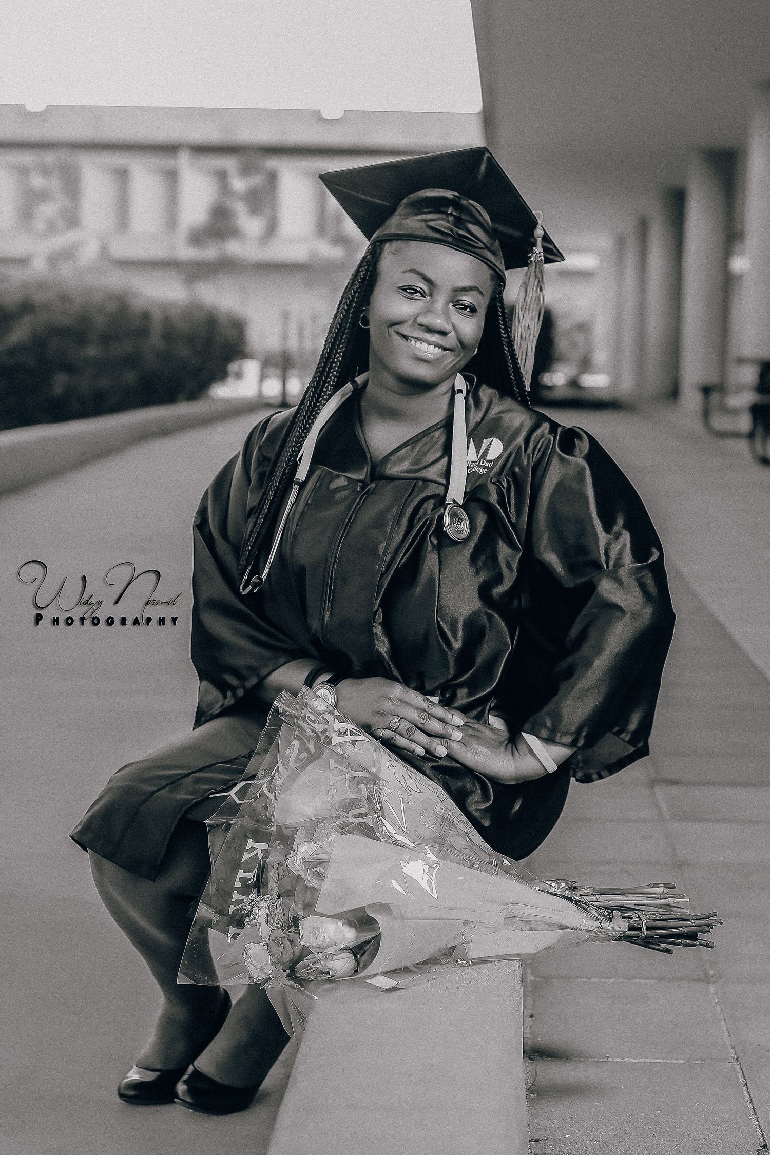Black and white portrait of a smiling graduate in cap and gown holding a bouquet of flowers.