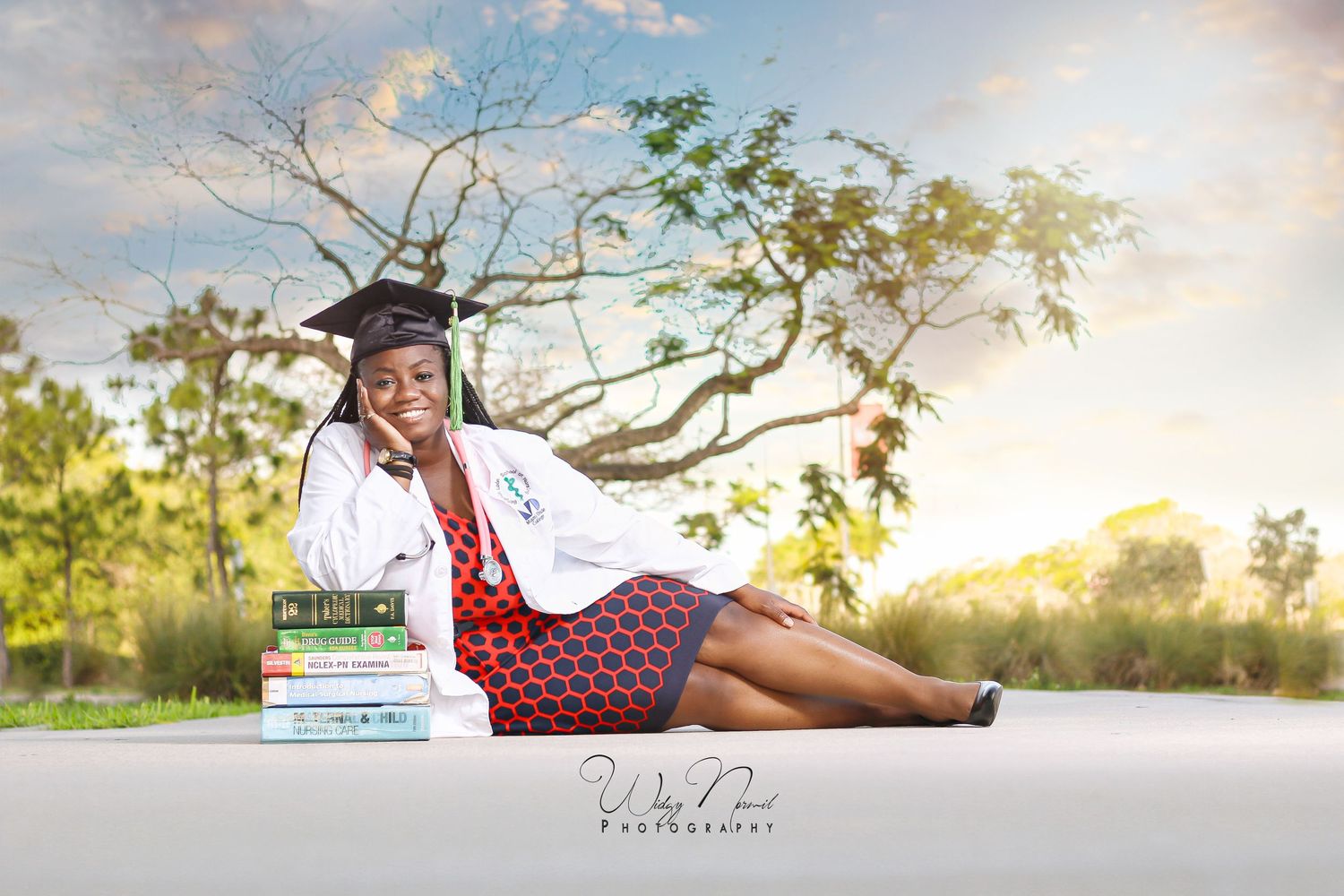 Smiling graduate in white coat and cap sits outdoors beside stacked books under a scenic sky.