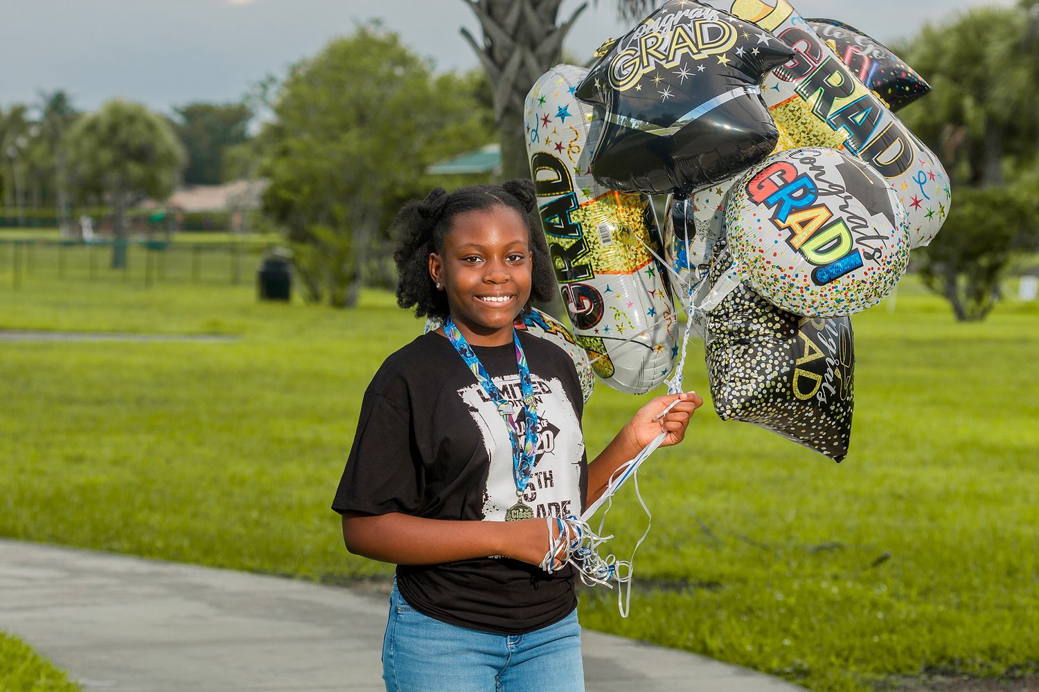 Smiling young Black girl holds colorful Congrats Grad balloons outdoors on green grass.