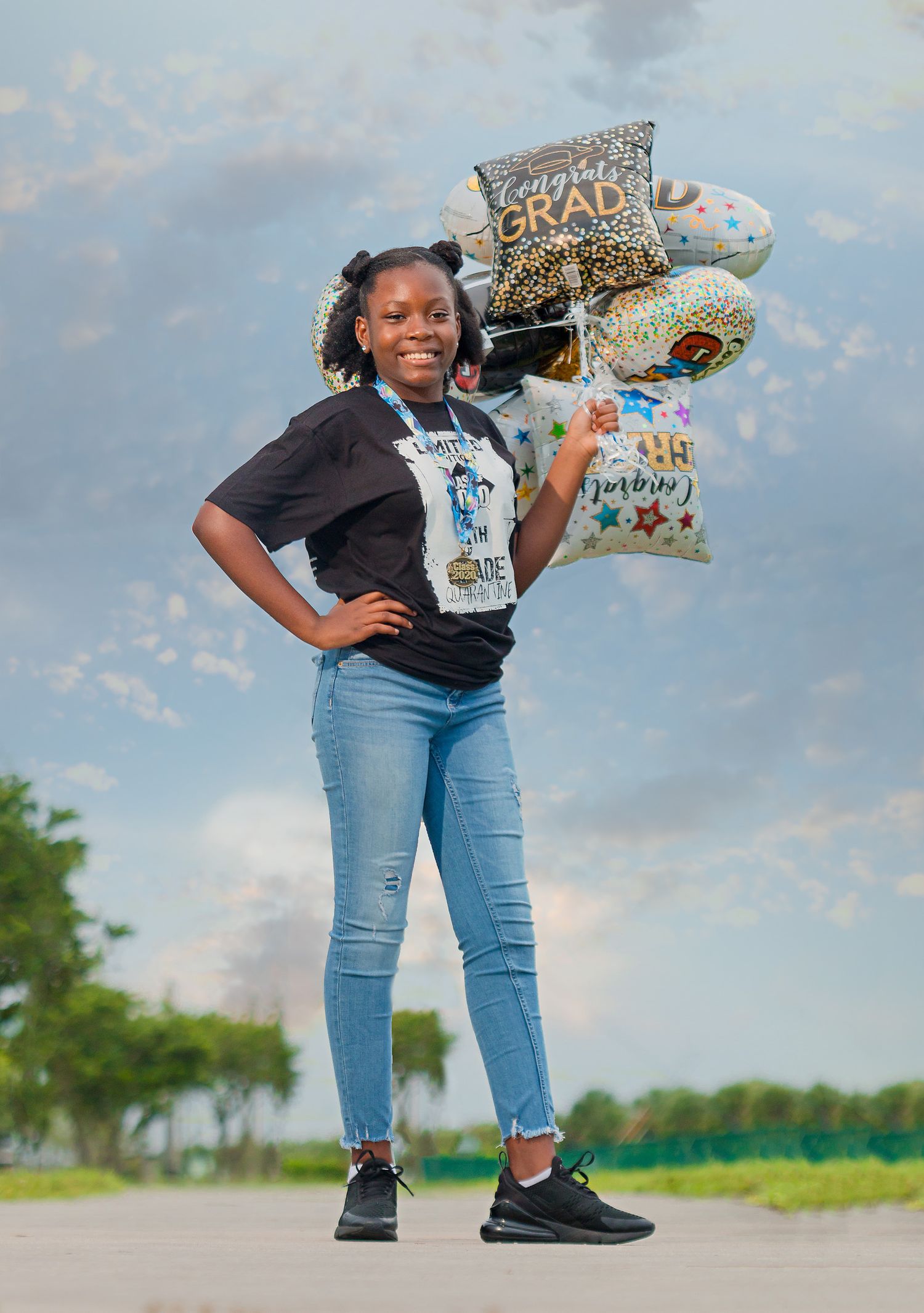 Young Black girl poses confidently with grad balloons wearing Class of 2020 5th grade shirt.
