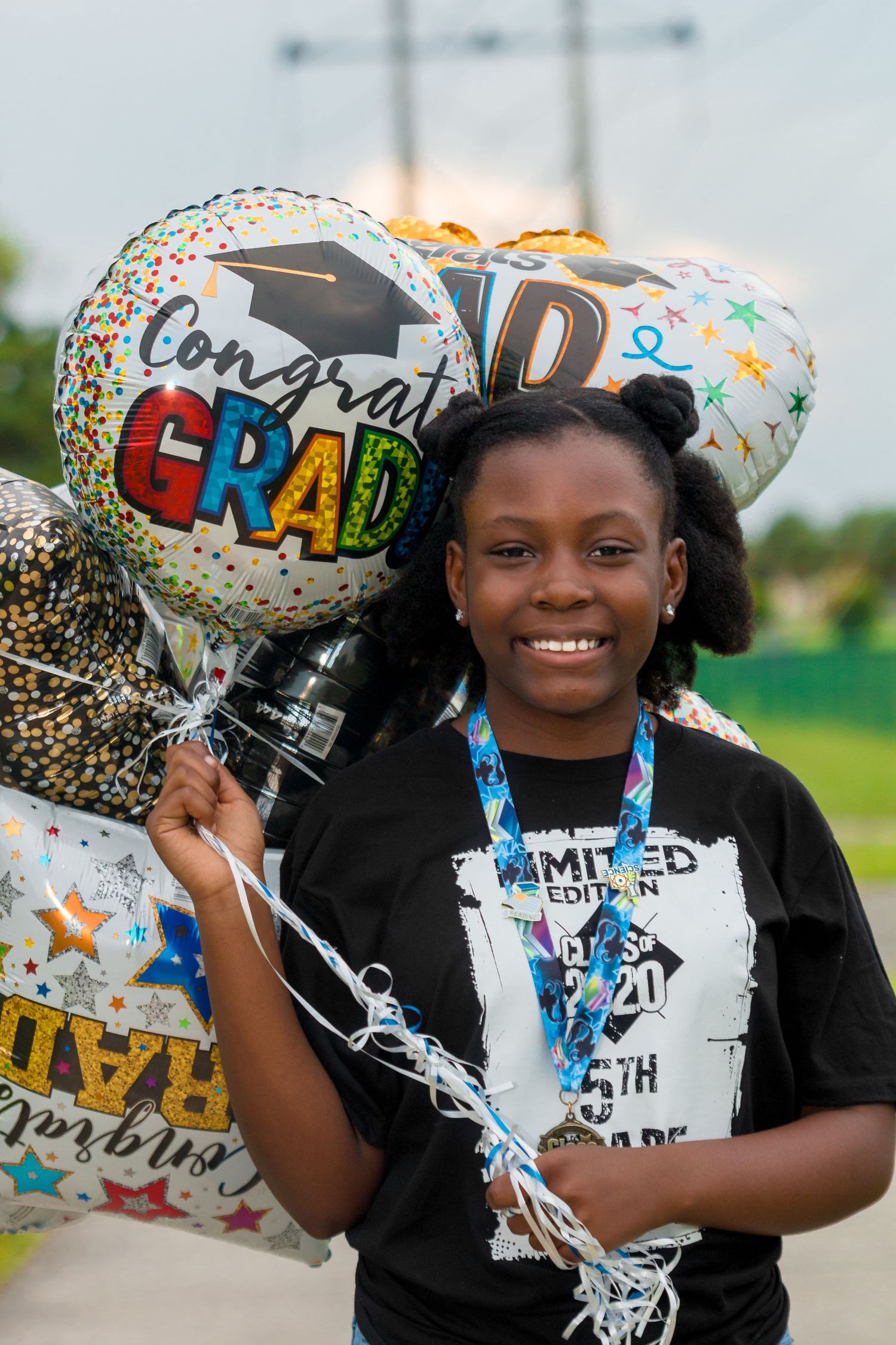 Smiling young Black girl holds Congrats Grad balloons wearing Class of 2020 5th grade shirt.