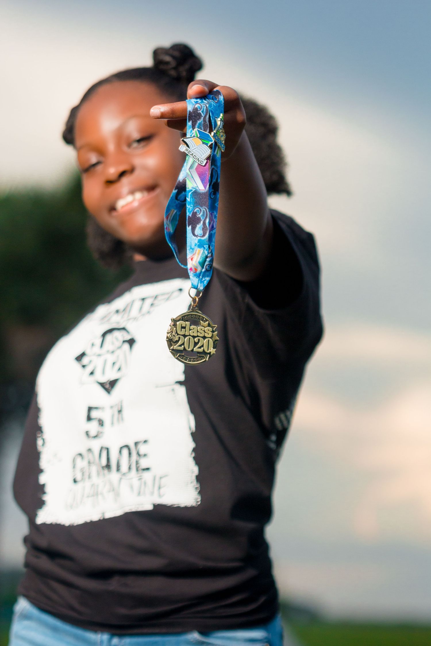 Young Black girl proudly displays Class of 2020 graduation medal toward camera outdoors.