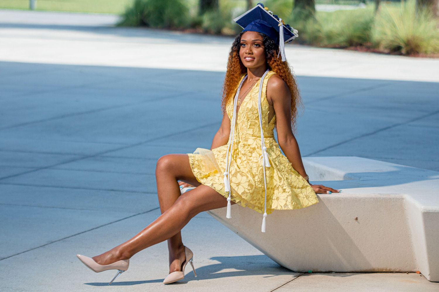 Female graduate in yellow dress and blue cap sits on outdoor bench, posing for senior photos.