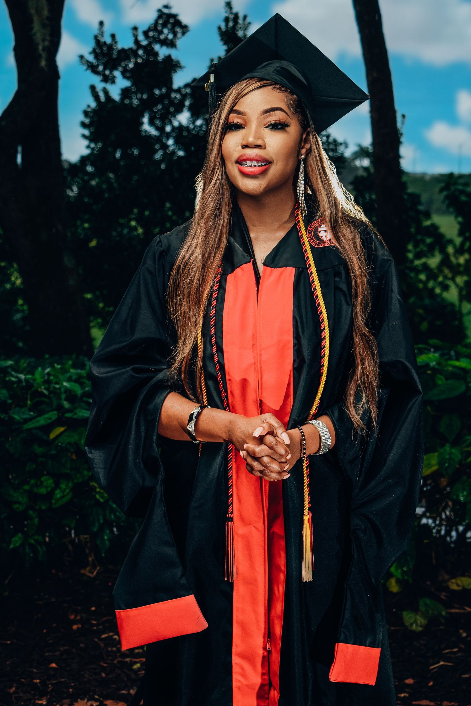 Female graduate in black and red cap and gown with honor cords stands outdoors among green foliage.