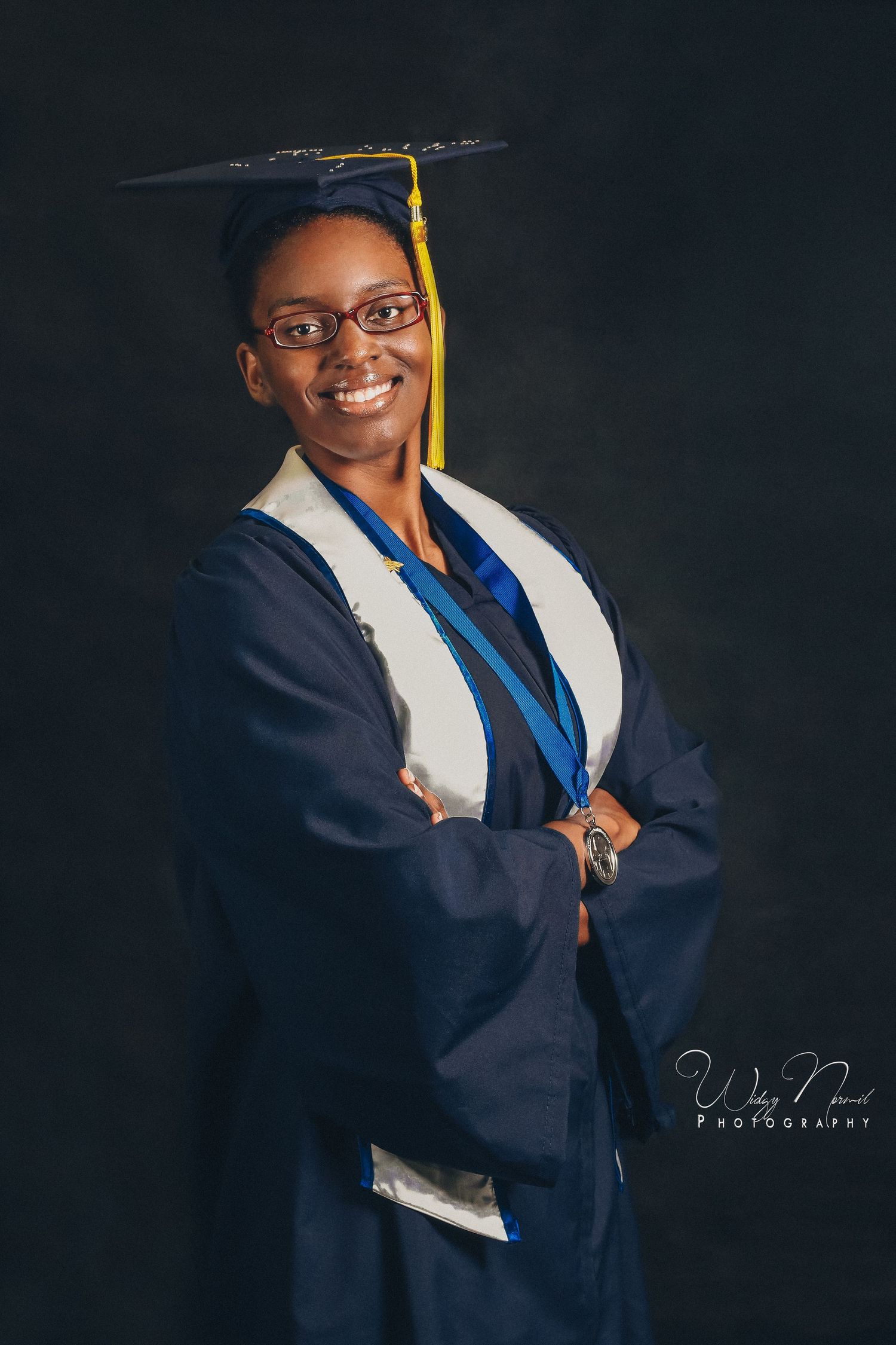 Proud female graduate in navy cap and gown stands with arms crossed, smiling for portrait photo.