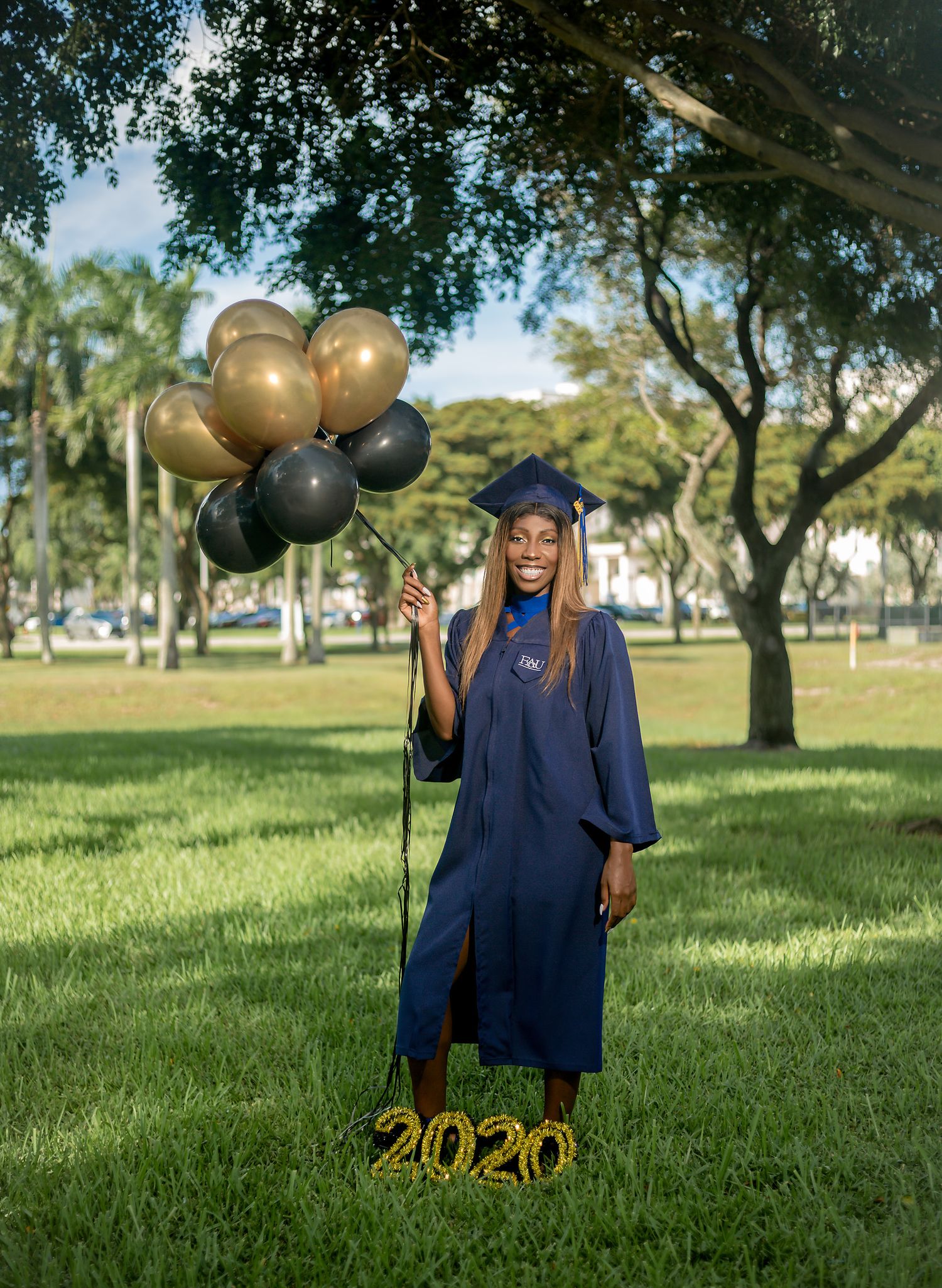 Smiling graduate in blue cap and gown holding gold and black balloons beside a 2020 sign outdoors.