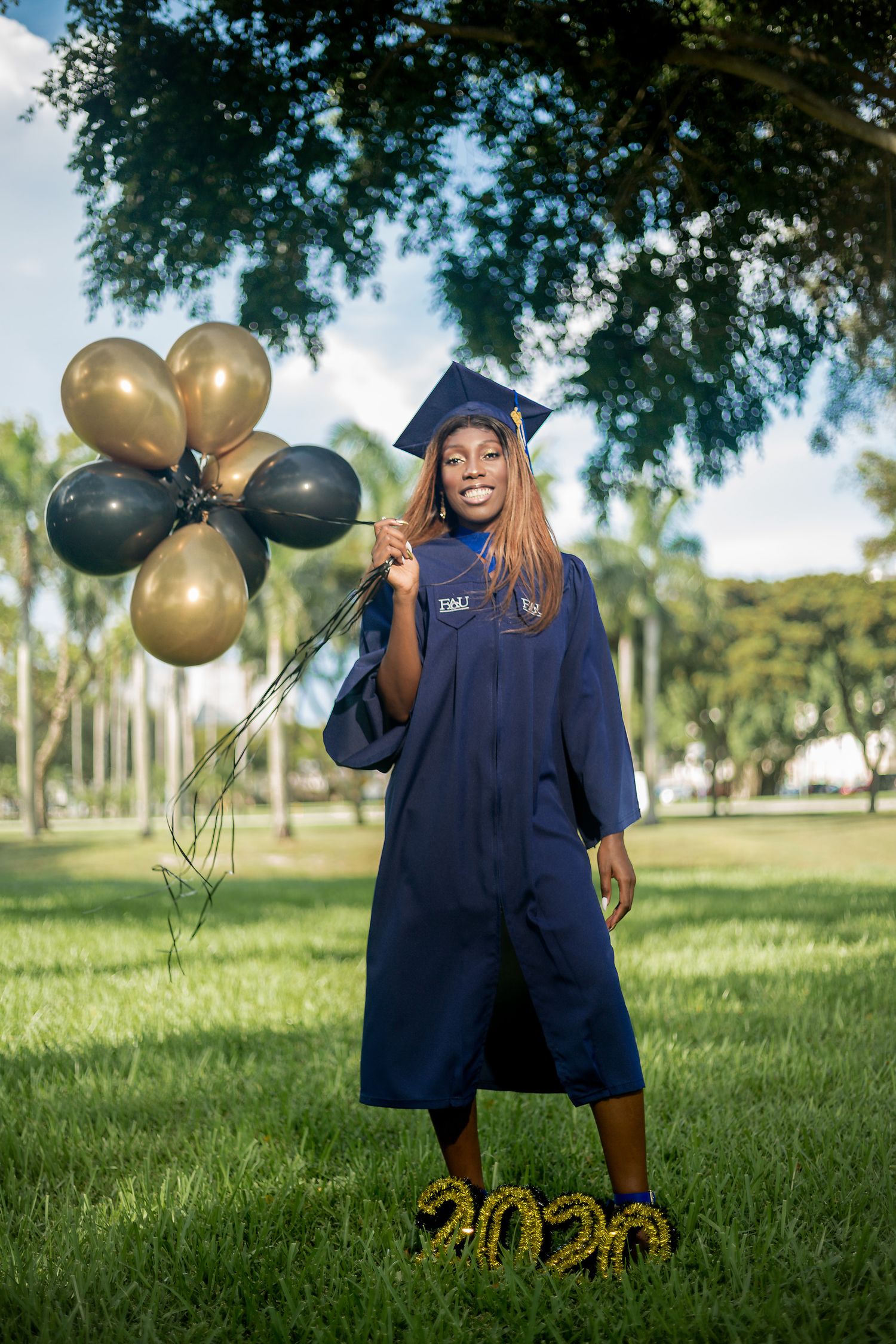 Female graduate in navy cap and gown holds gold and black balloons near 2020 sign on green lawn.
