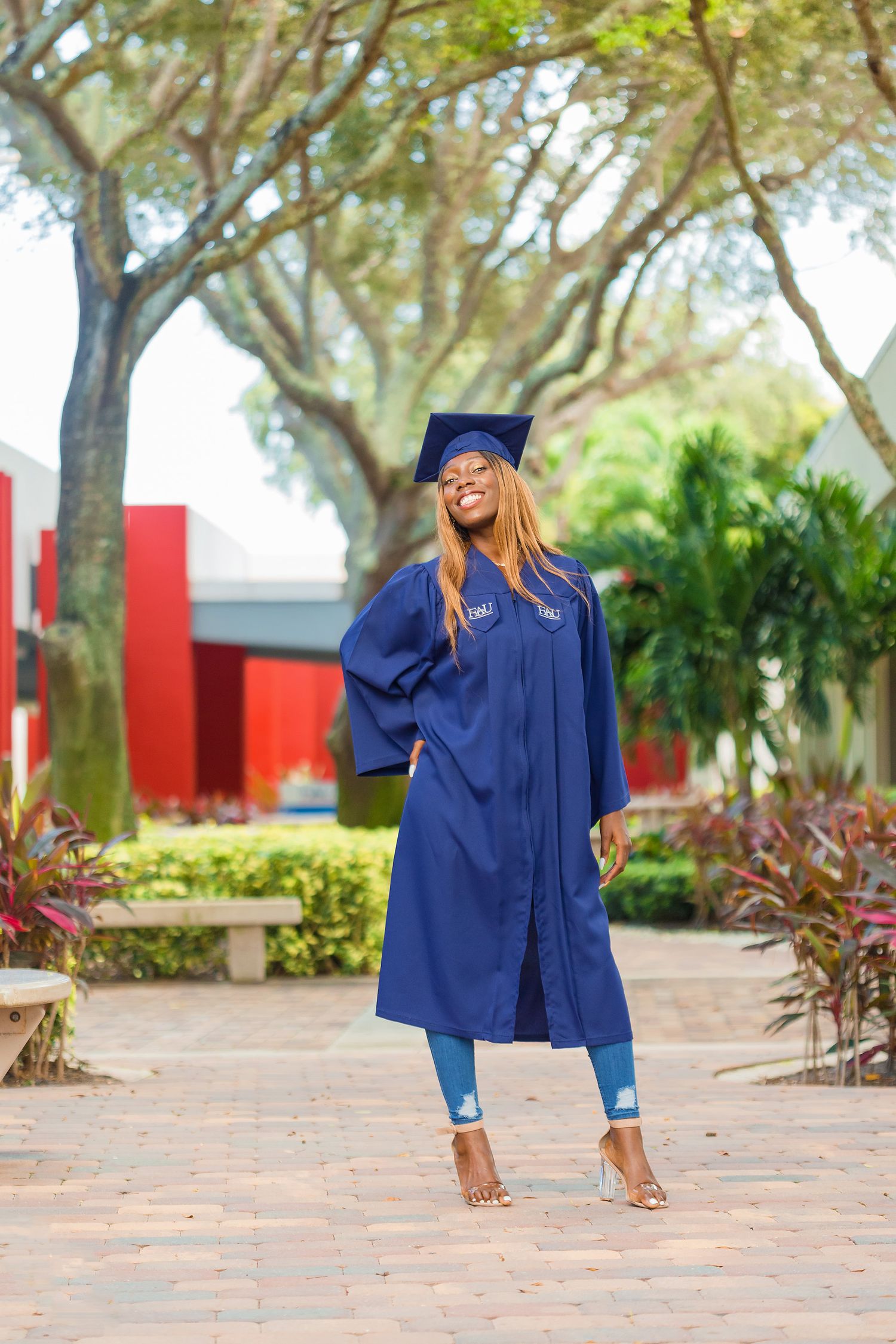 Graduate in blue cap and gown posing confidently on a tropical university campus pathway.