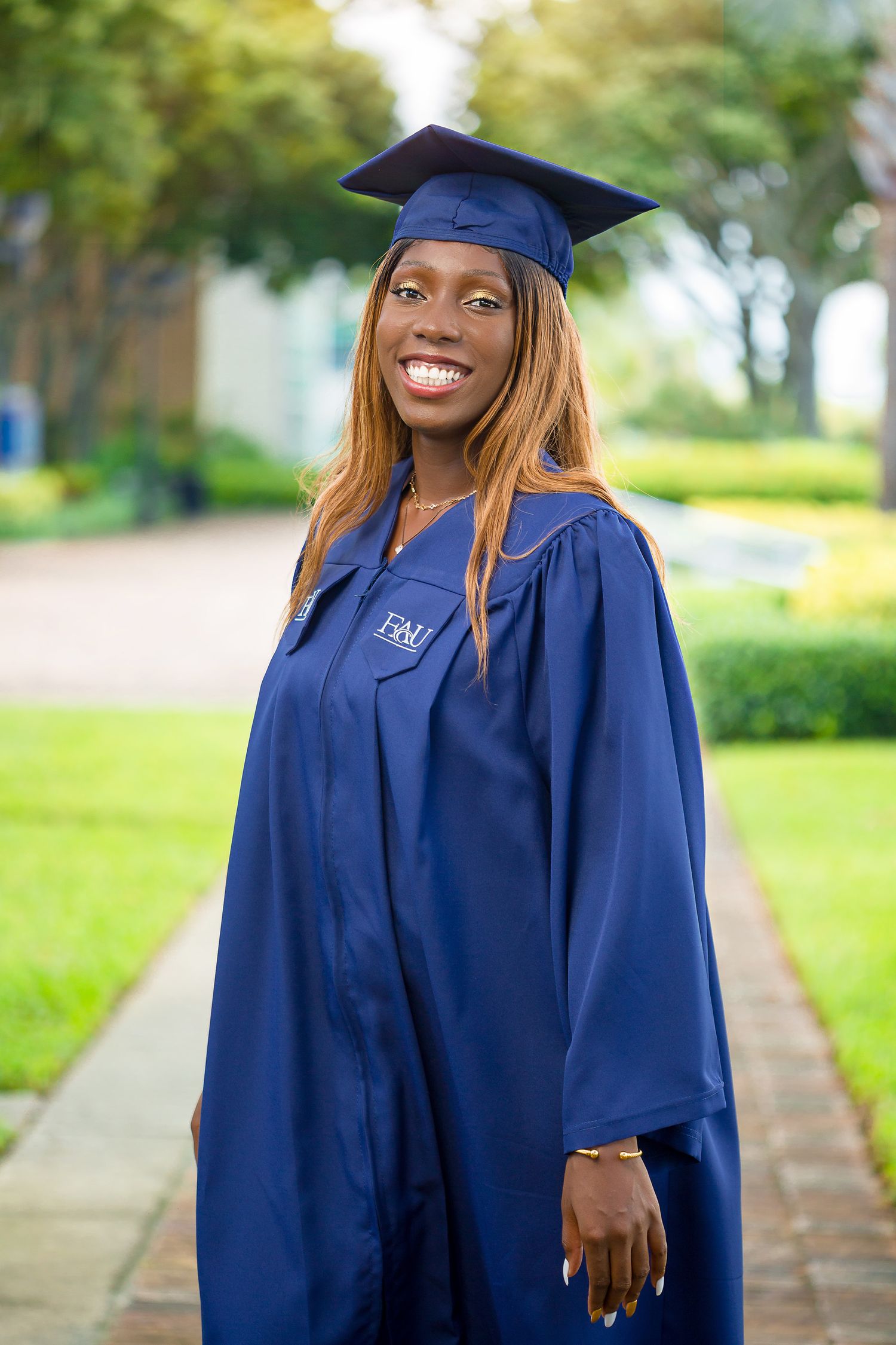 Young woman in navy blue FIU graduation cap and gown smiling on a sunny campus walkway.