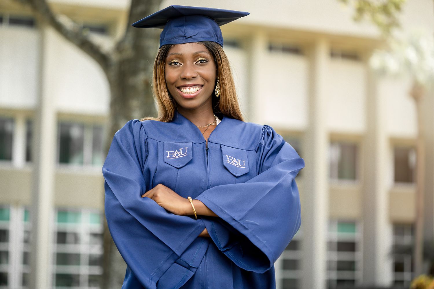 Smiling graduate in blue cap and gown with arms crossed standing in front of a campus building.