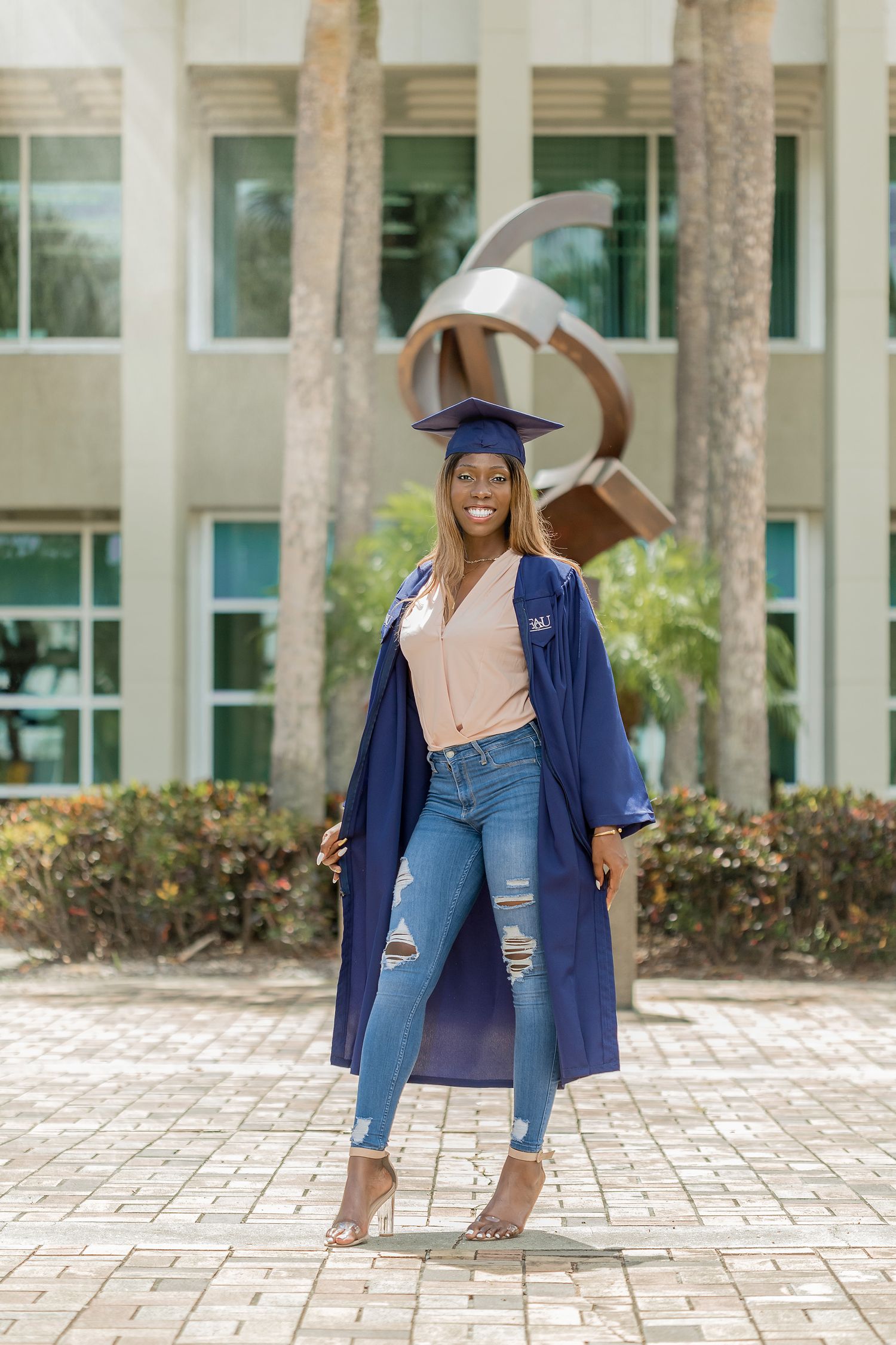 Graduate in open blue gown and ripped jeans posing before a modern sculpture on campus.