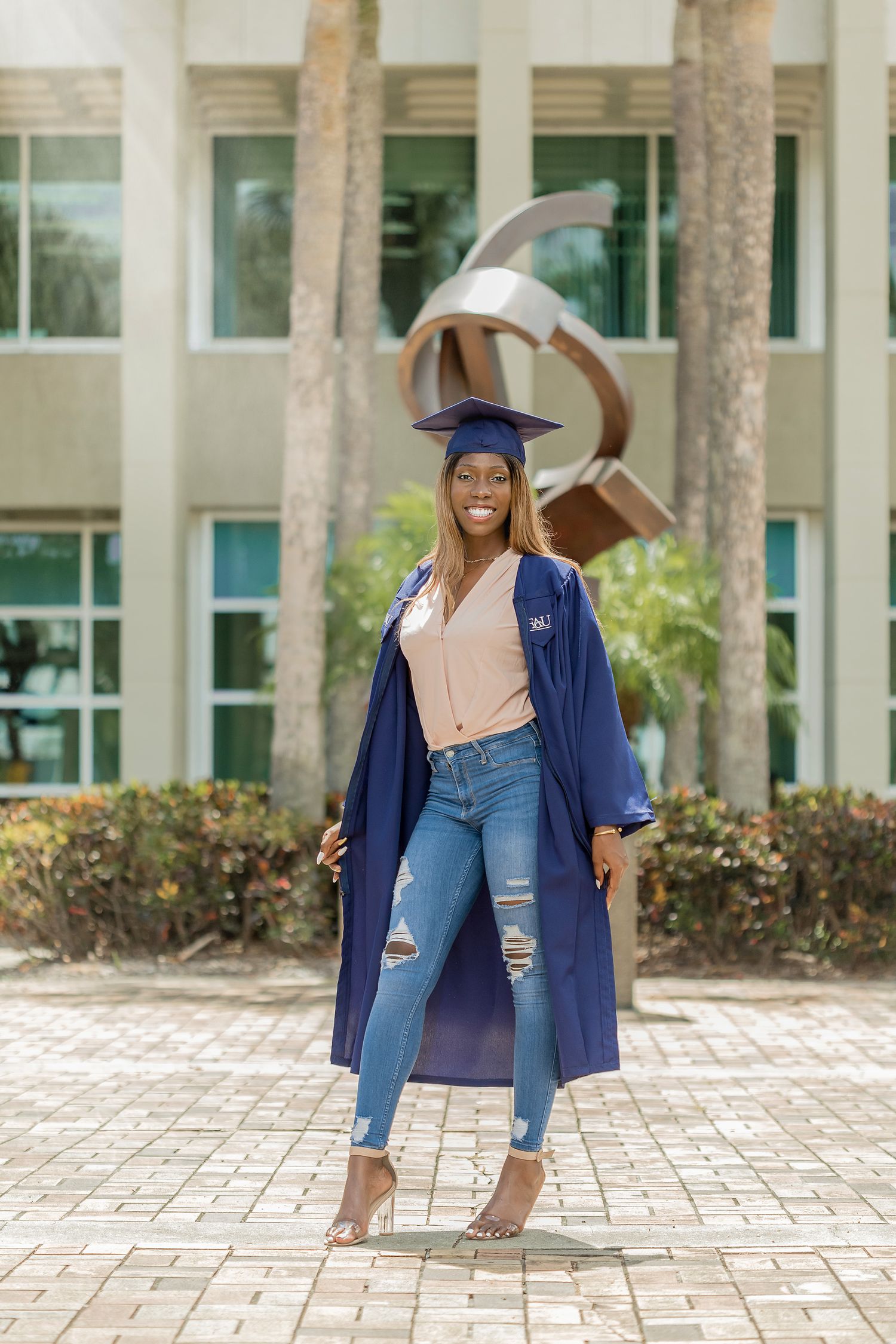 Smiling graduate in blue cap and open gown with ripped jeans standing by a campus sculpture.