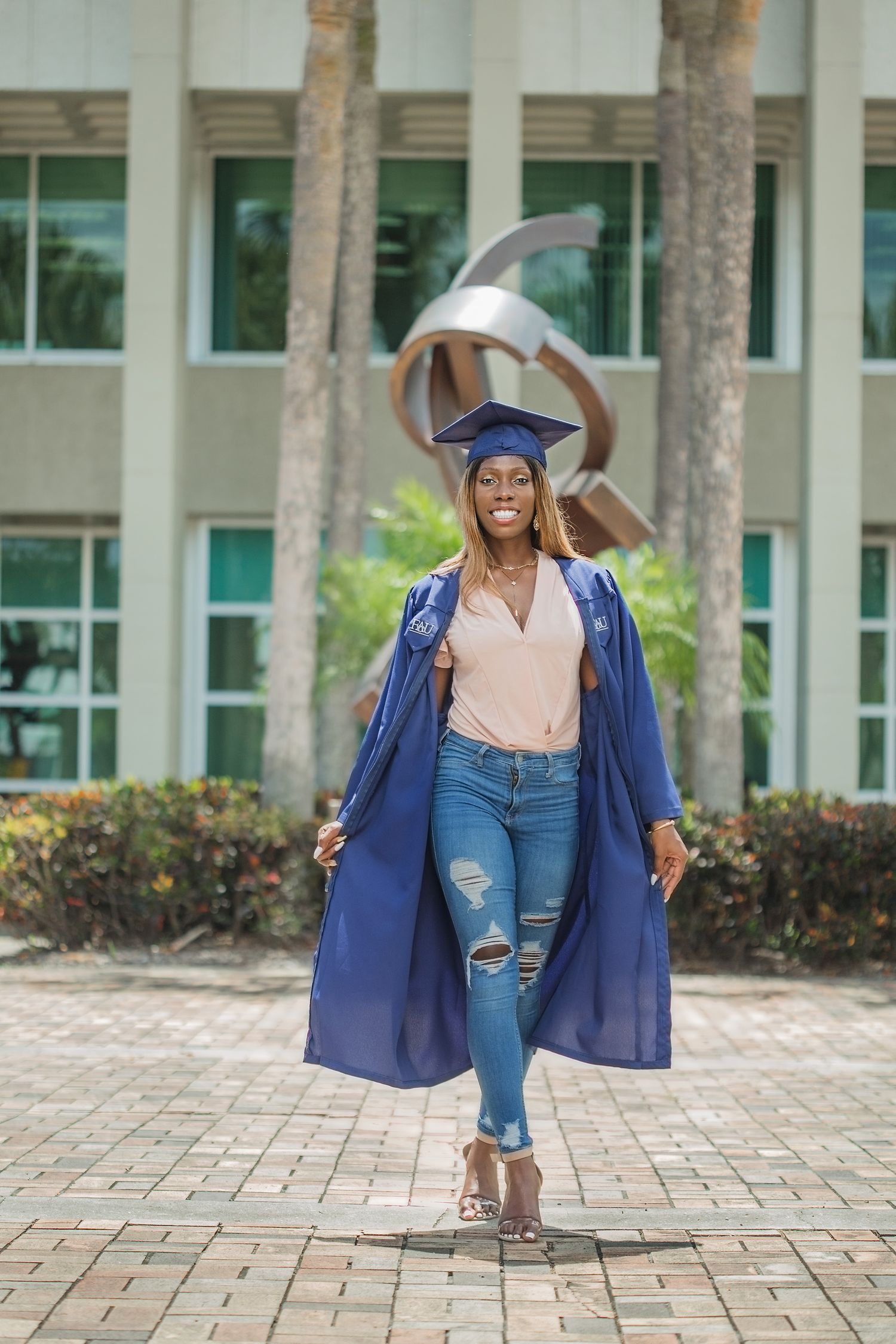Joyful graduate in blue cap and gown walking confidently in front of a campus sculpture.