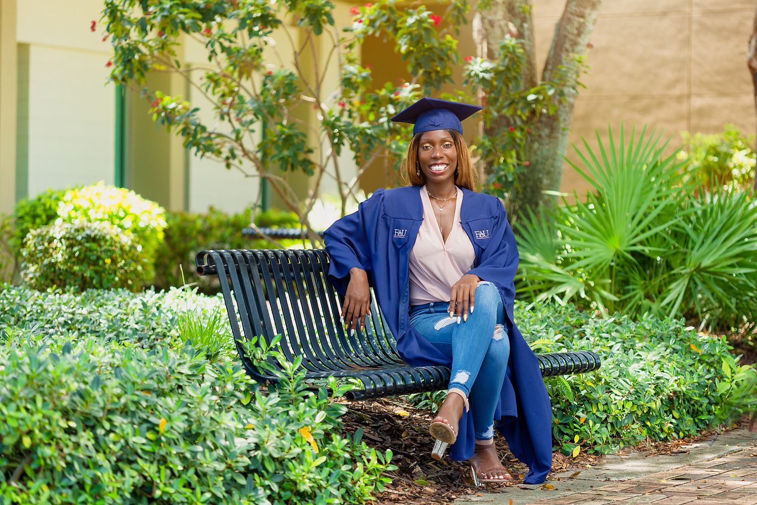 Graduate in blue cap and gown sitting on a bench surrounded by lush greenery on campus.