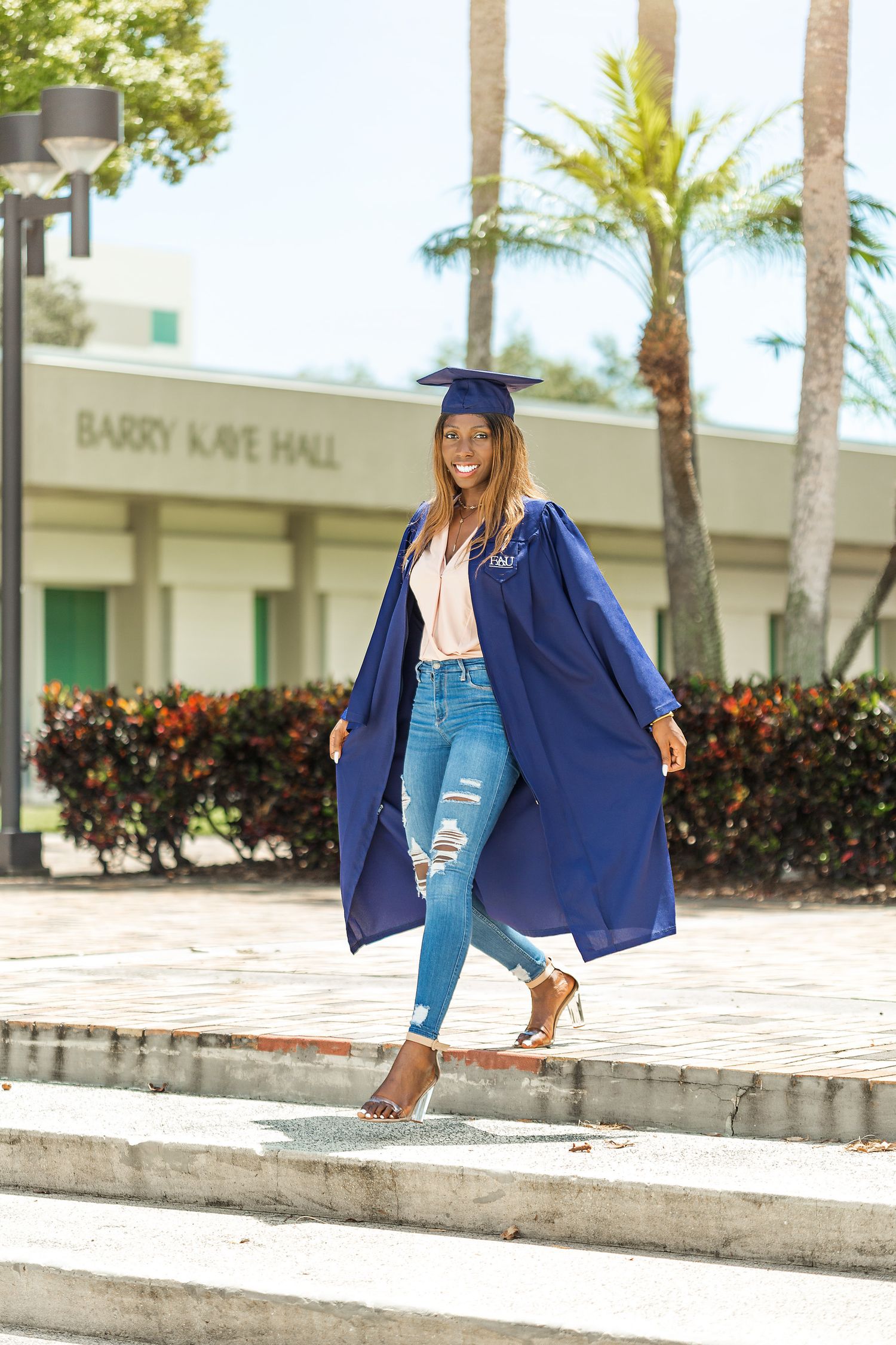 Smiling Black woman in blue graduation cap and gown poses on campus steps at Barry Kaye Hall.