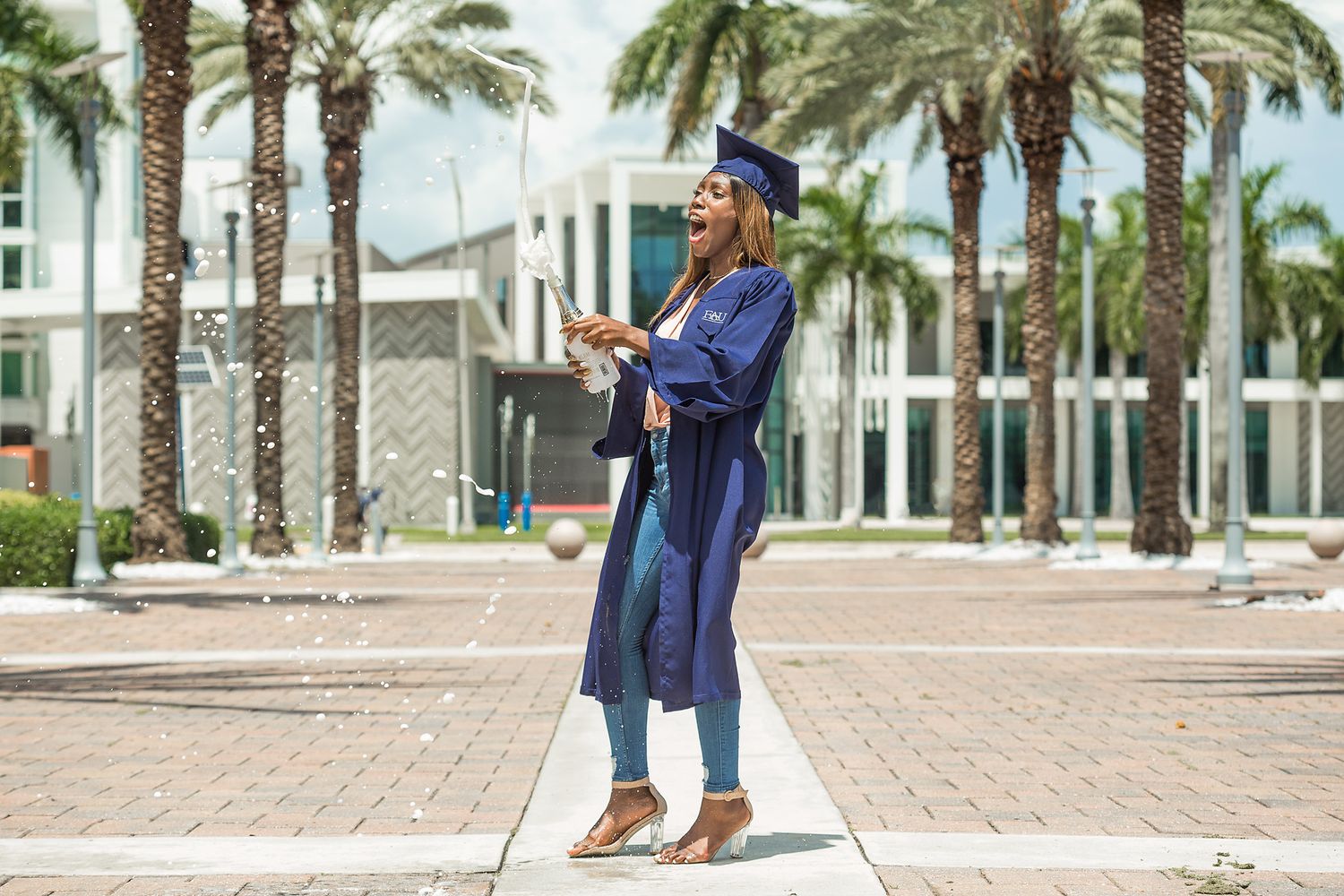 Graduate in blue cap and gown tossing confetti near palm trees outside a modern building.