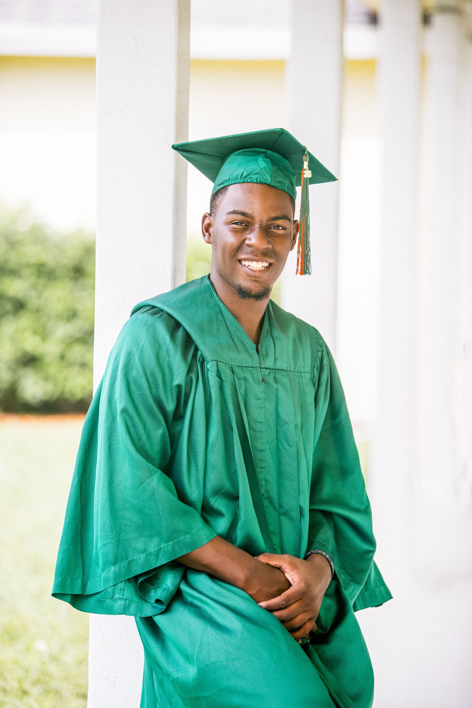 Smiling young man in green graduation cap and gown poses confidently between white columns outdoors.