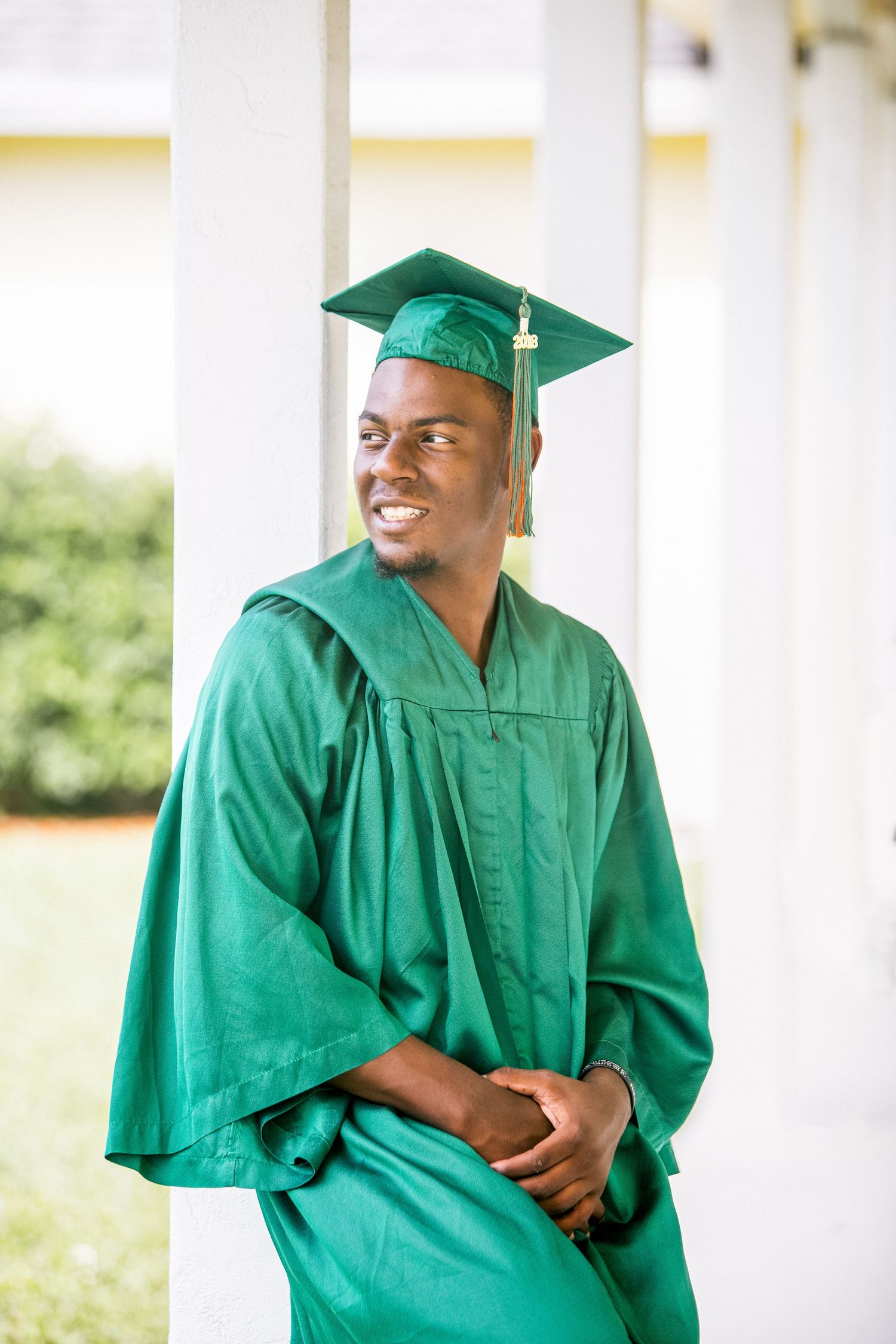 Young male graduate in green cap and gown leans against white column, smiling and looking away.