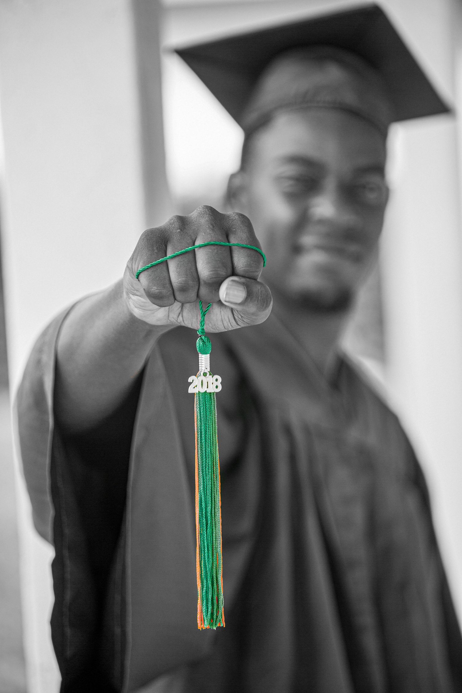 Graduate holds up green and gold 2018 graduation tassel in black and white selective color photo.