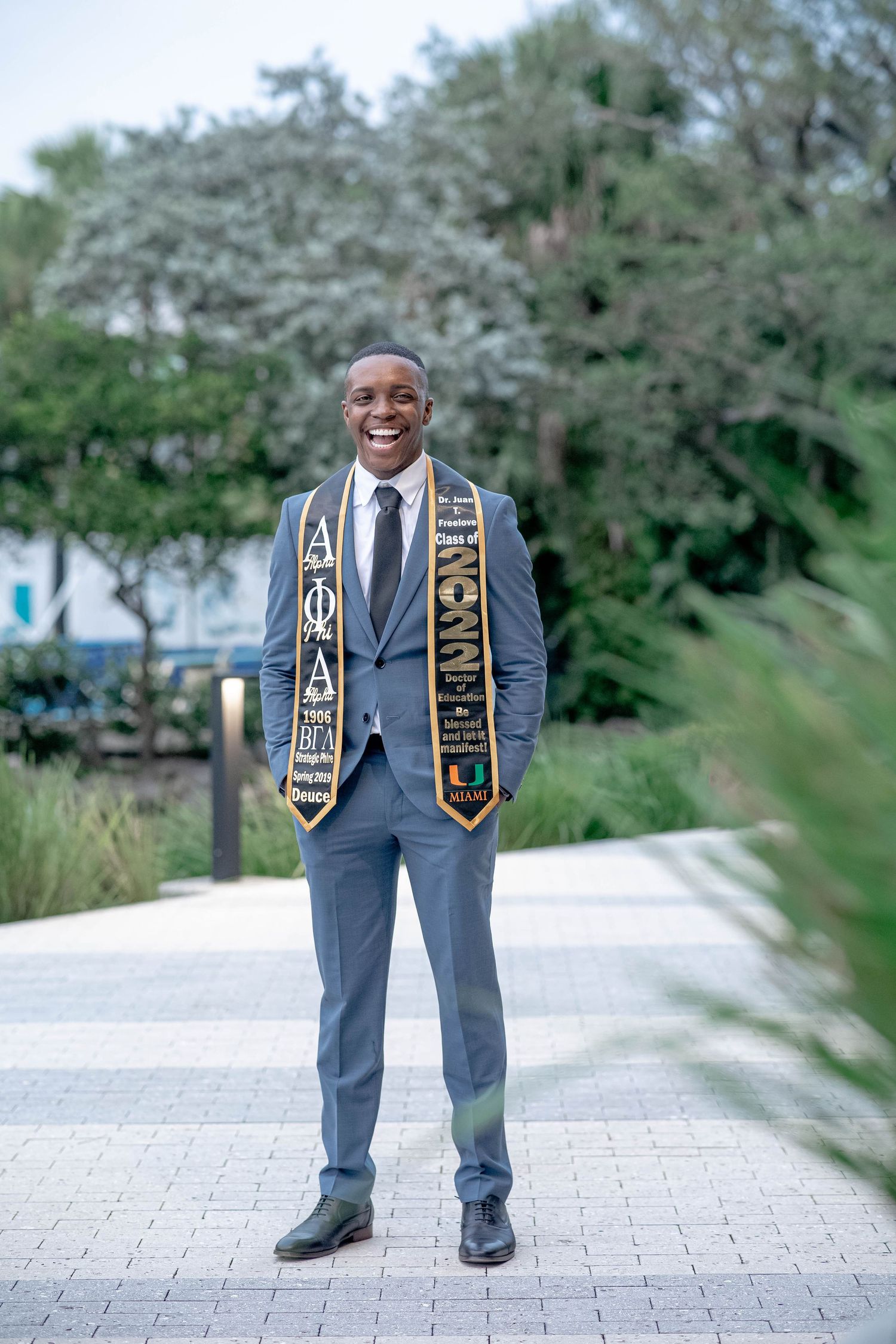 Smiling Black male graduate stands on walkway wearing University of Miami Alpha Phi Alpha stole.