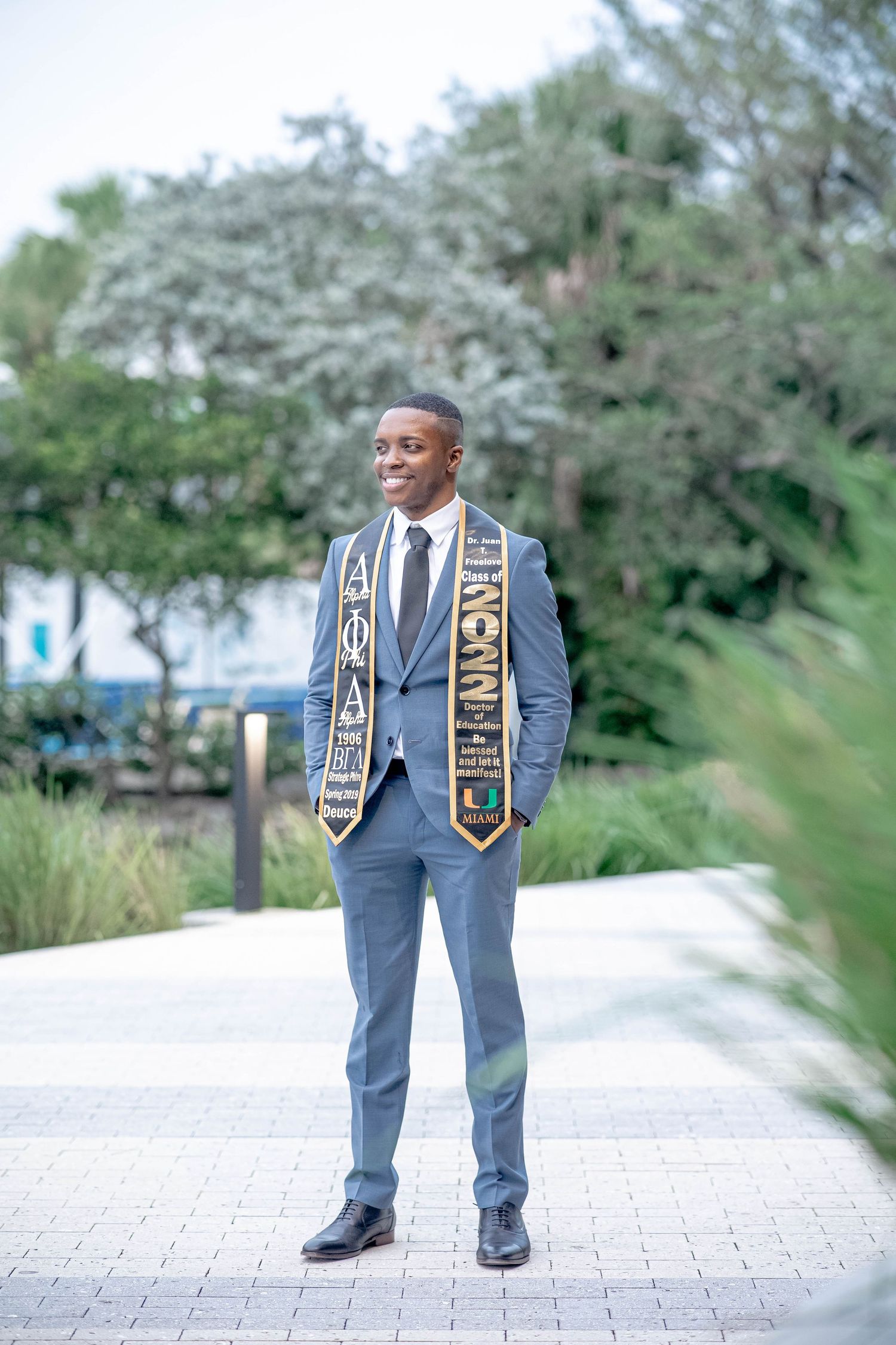 Young man in gray suit wearing graduation stoles smiles outdoors on brick pathway with green trees behind him.