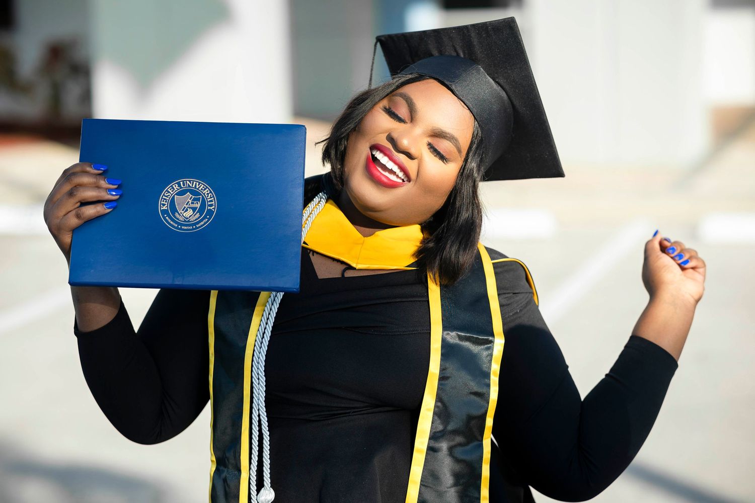 Joyful graduate in black cap and gown celebrating while holding a blue diploma outdoors.